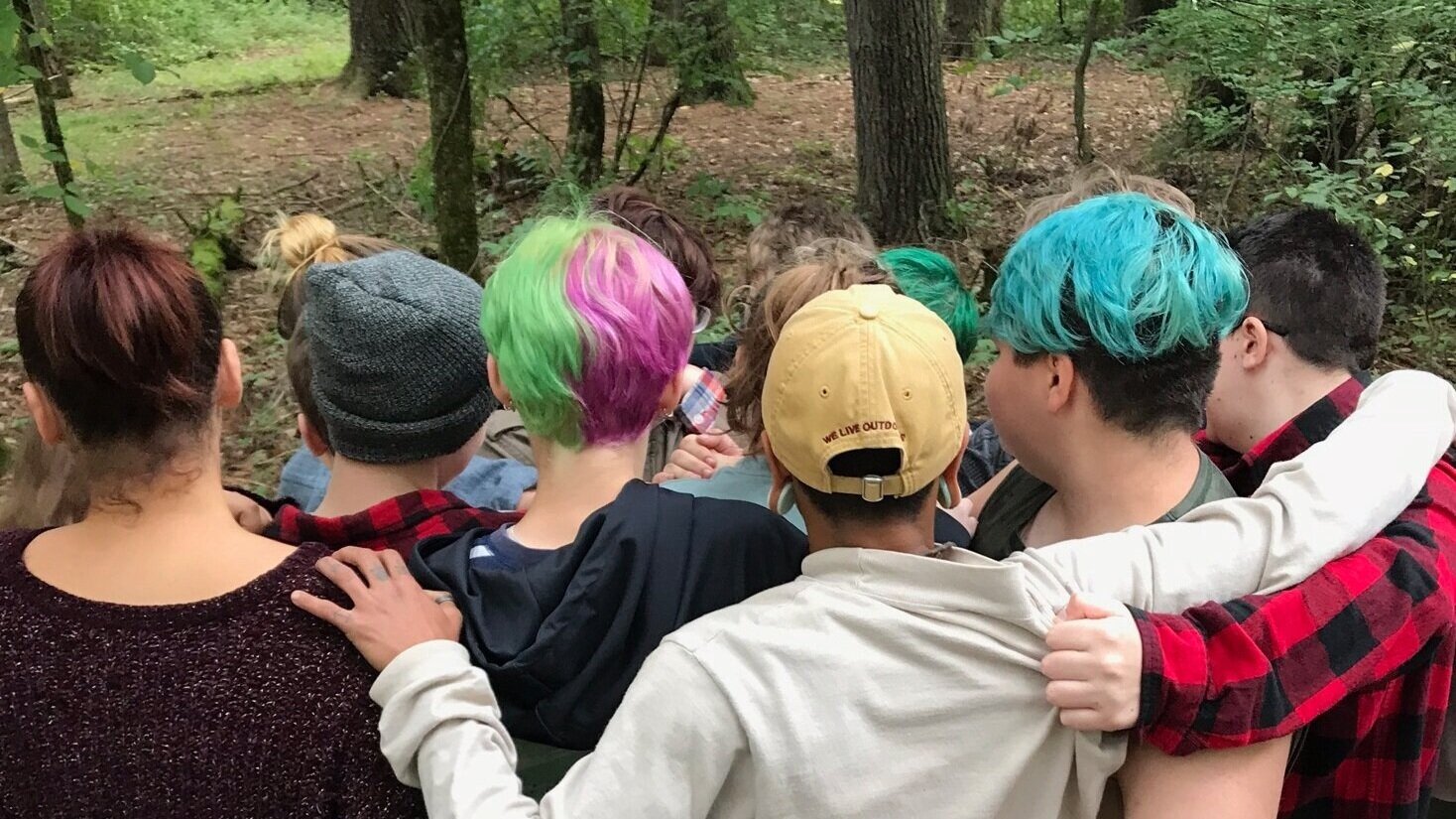  A group of seven teens are clustered together facing away from the camera. One individual is seen facing the camera between the shoulders of the teens. This individual is looking at their wrist. 