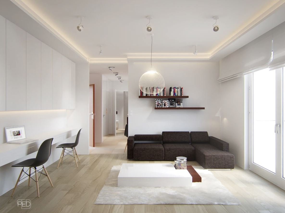Modern living room with a dark sectional sofa, white coffee table, built-in wall shelves with books and photos, large window with blinds, and a white wall with crown molding and lighting accents.