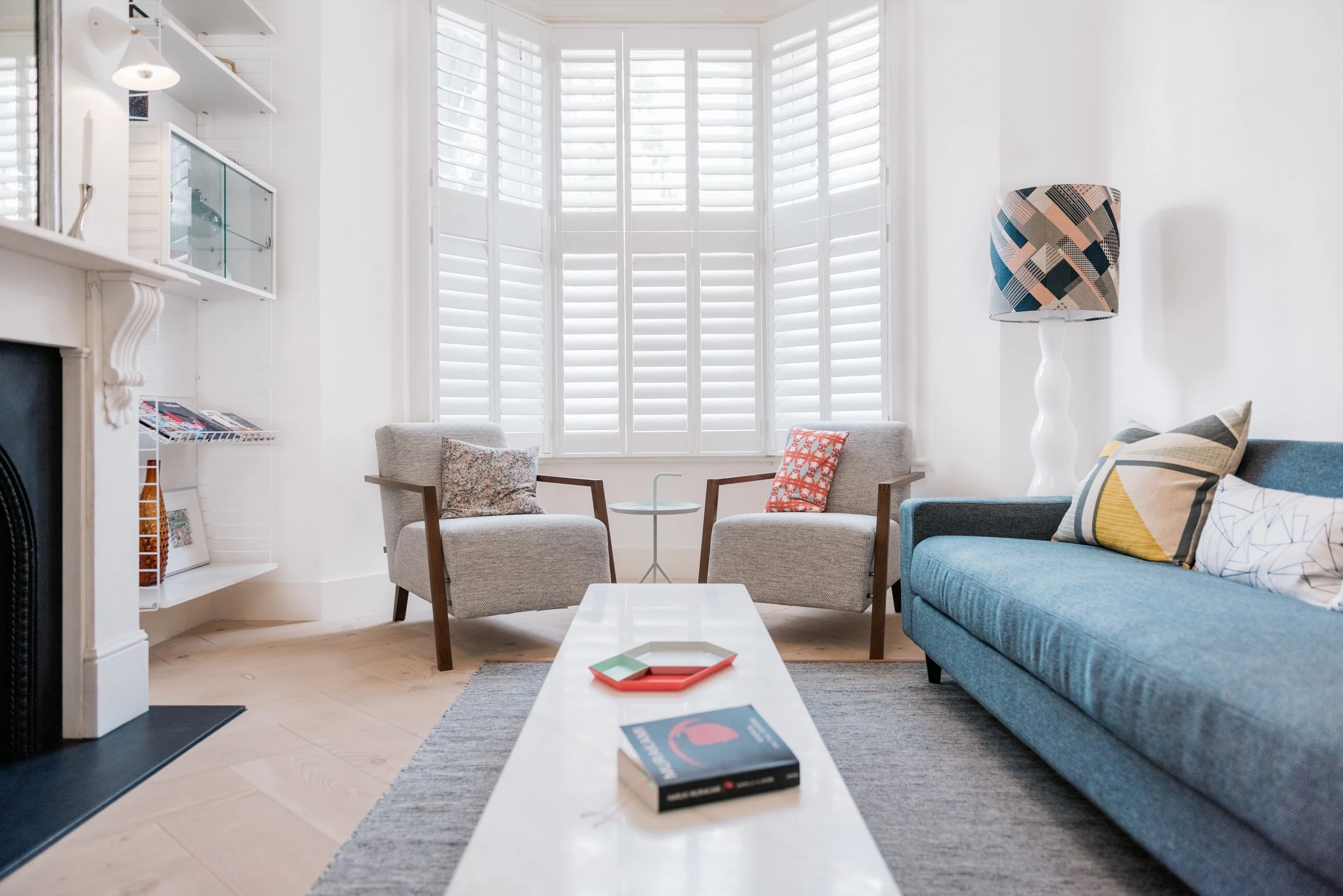 Bright living room with a large window with white shutters, gray and blue sofas, patterned cushions, a white coffee table, and a floor lamp with a patterned shade.
