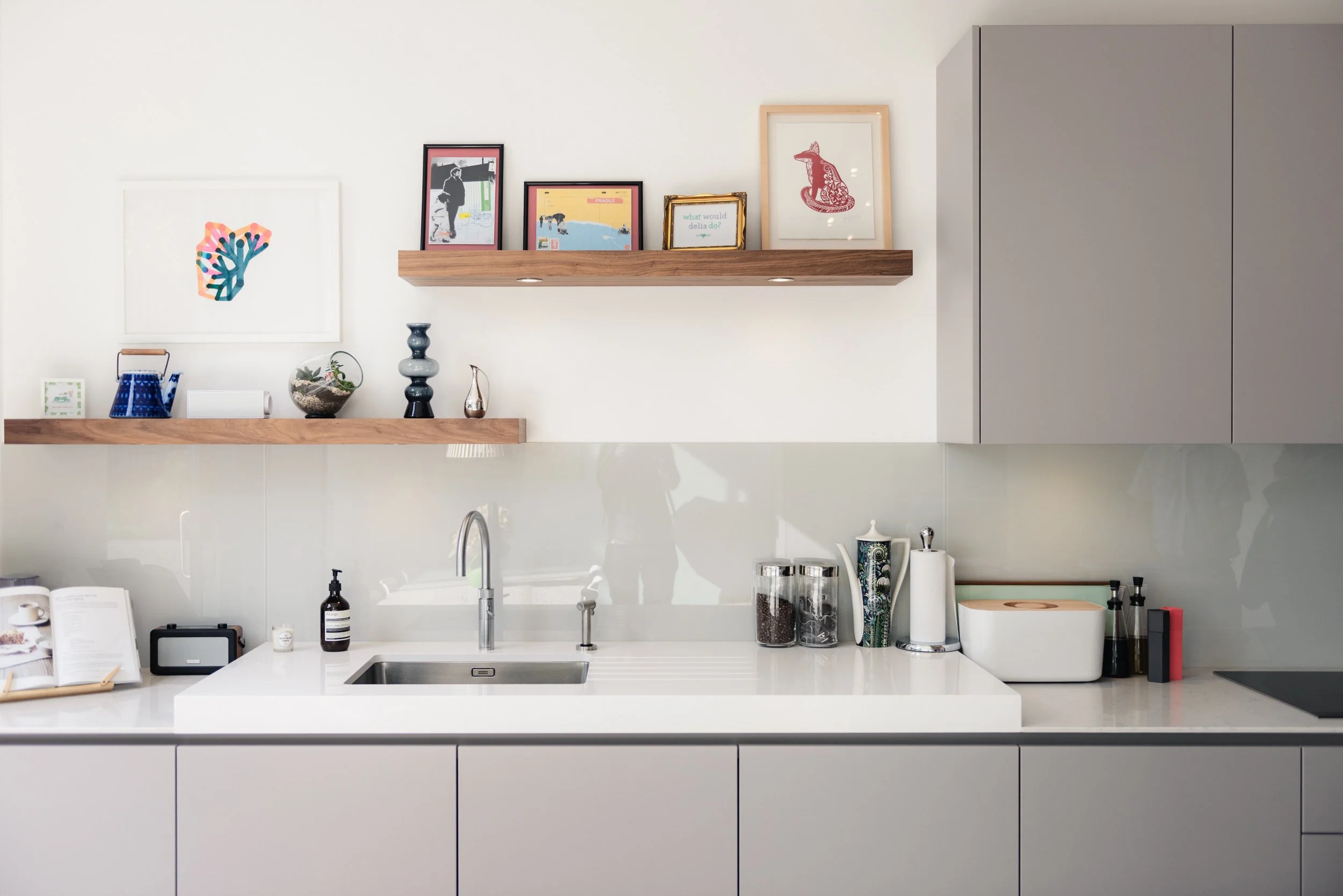 Modern kitchen with white cabinetry, a white countertop, a stainless steel sink, and a white backsplash. There are two floating wooden shelves with framed artwork and decorative items. The artwork includes colorful abstract and illustrated pieces. It