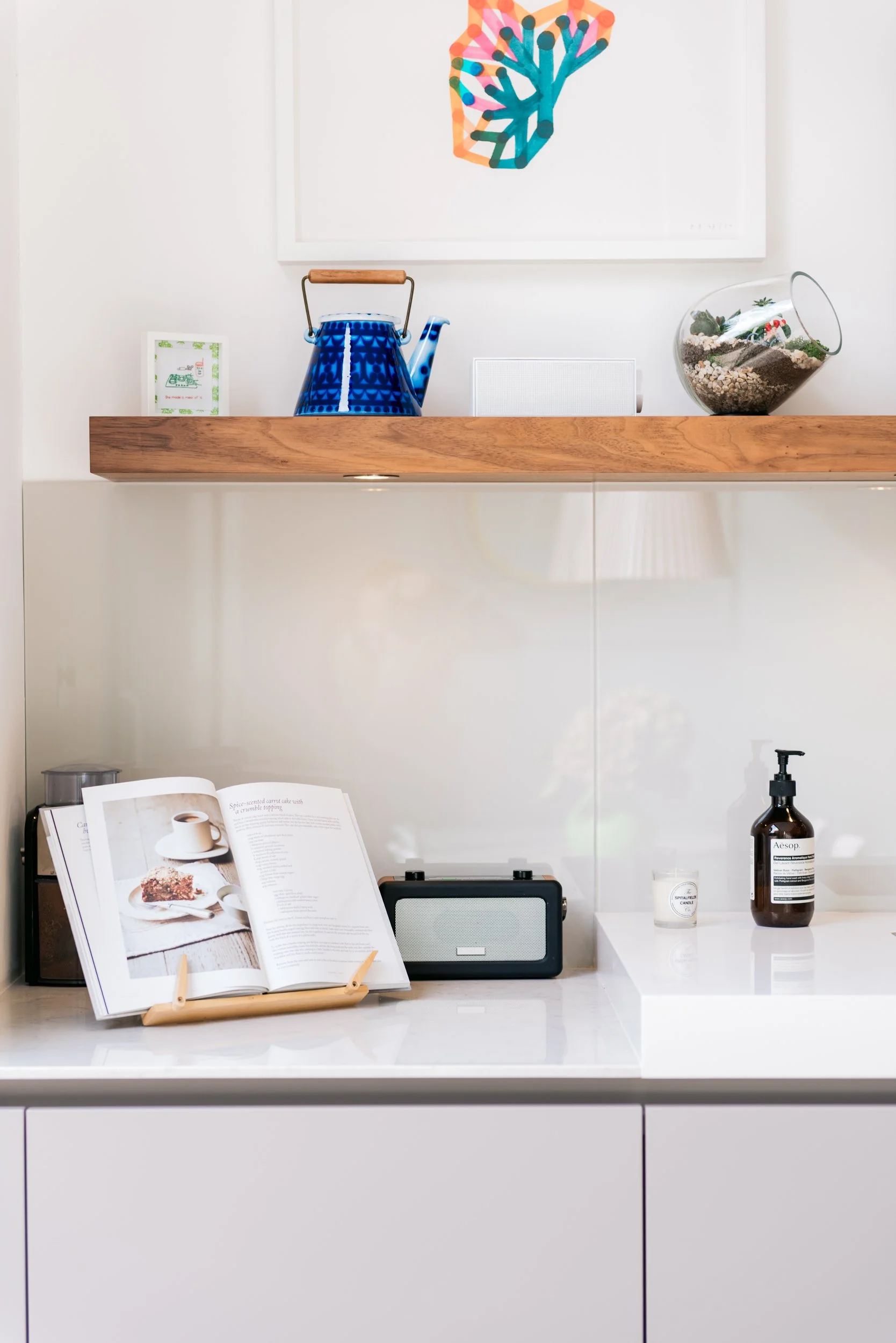 Kitchen shelf with a blue tea kettle, a glass terrarium with plants, a small framed picture, a speaker, an open cookbook on a wooden easel, a bottle of Aesop hand soap, a candle, and a white countertop.