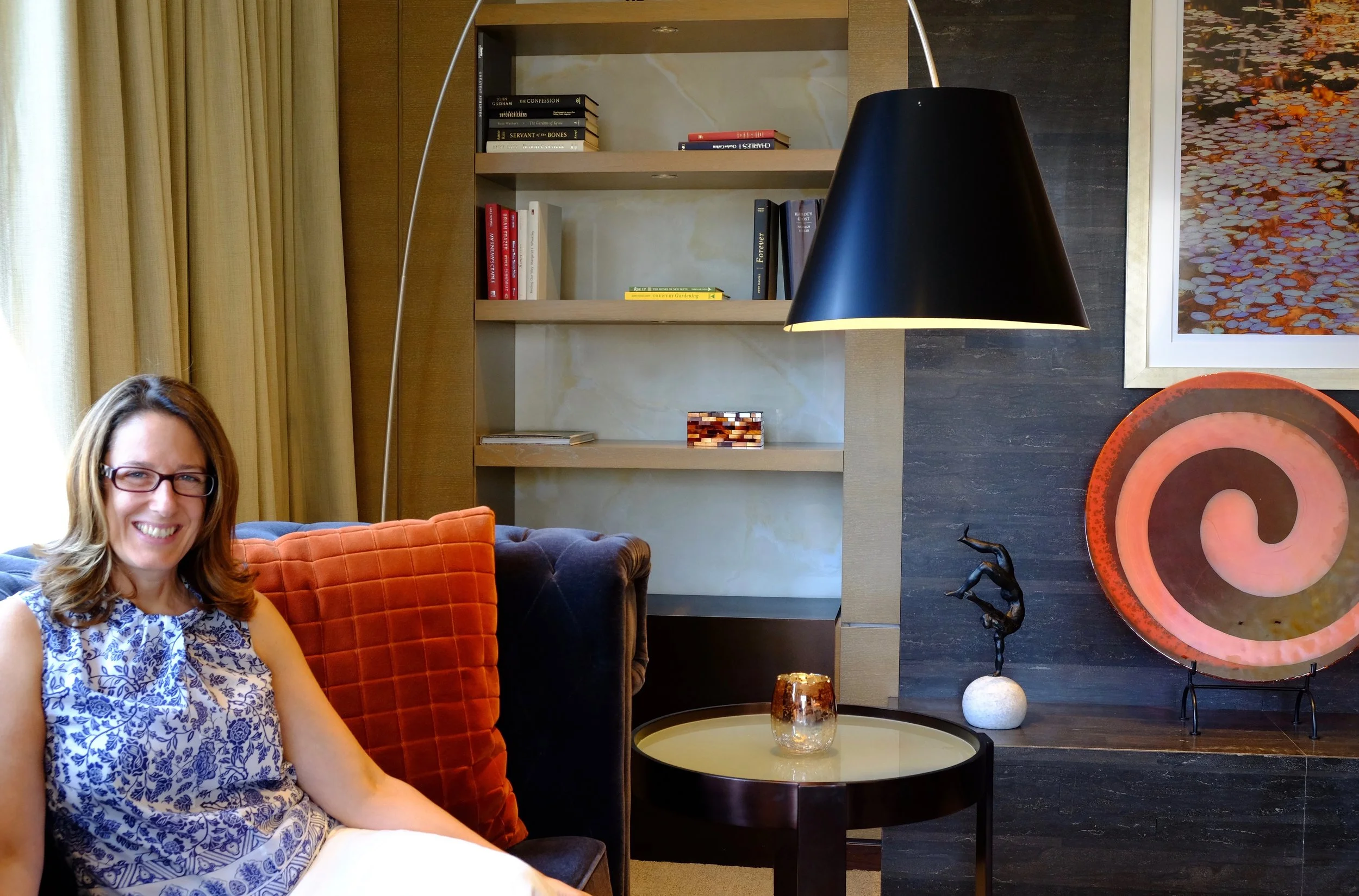 A woman with glasses and a blue patterned top sitting on a dark couch with orange and dark pillows, in a living room with a black lamp, bookshelf, wall art, and decorative objects.