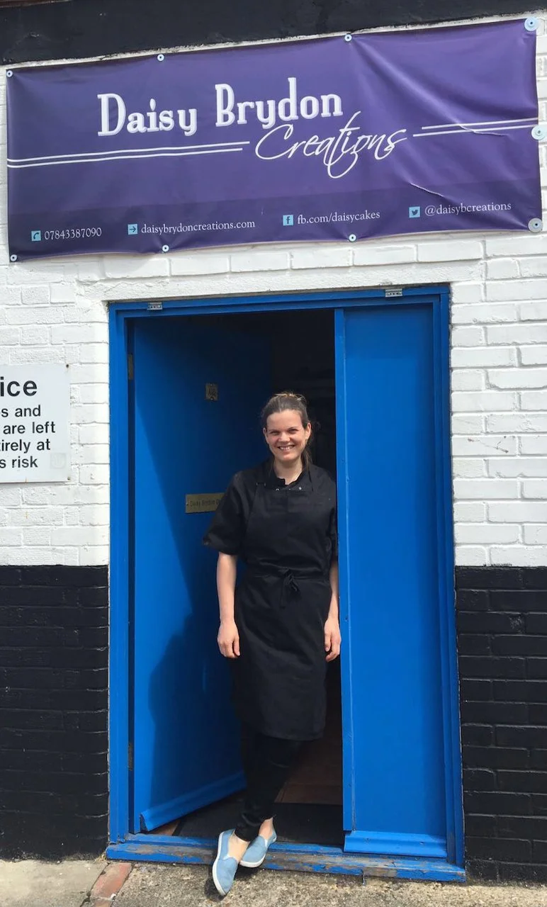 Smiling woman in black chef uniform standing by a blue door in a bakery or cake shop.