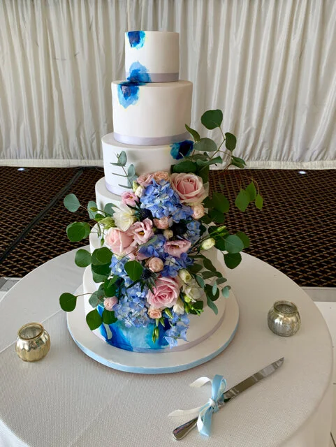 6 tier fondant wedding cake with blue accents decorated in pink and blue fresh fresh flowers and green foliage