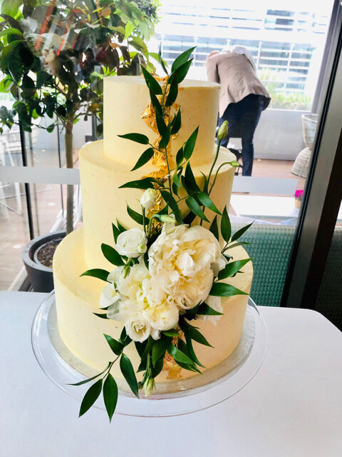 3 tier buttercream wedding cake with gold accents down the centre decorated with fresh flowers and foliage