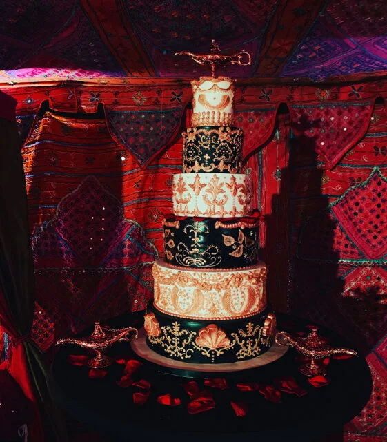 A luxury Arabian Nights themed 6 tier wedding cake in black, white and gold topped with a genie lamp