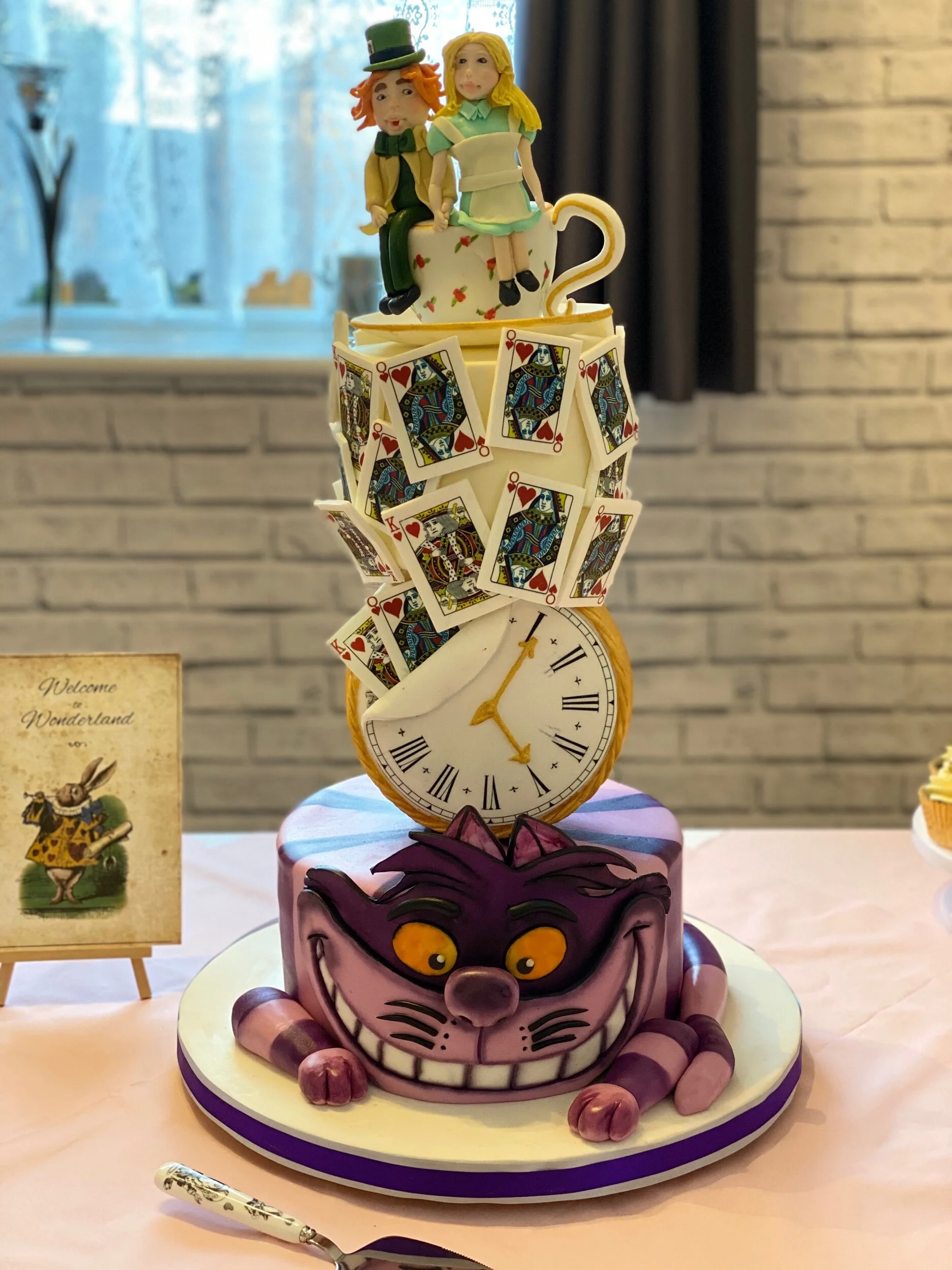 An Alice in Wonderland themed wedding cake. The bottom tier is the Cheshire cat. The tiers are separated with a working clock cake