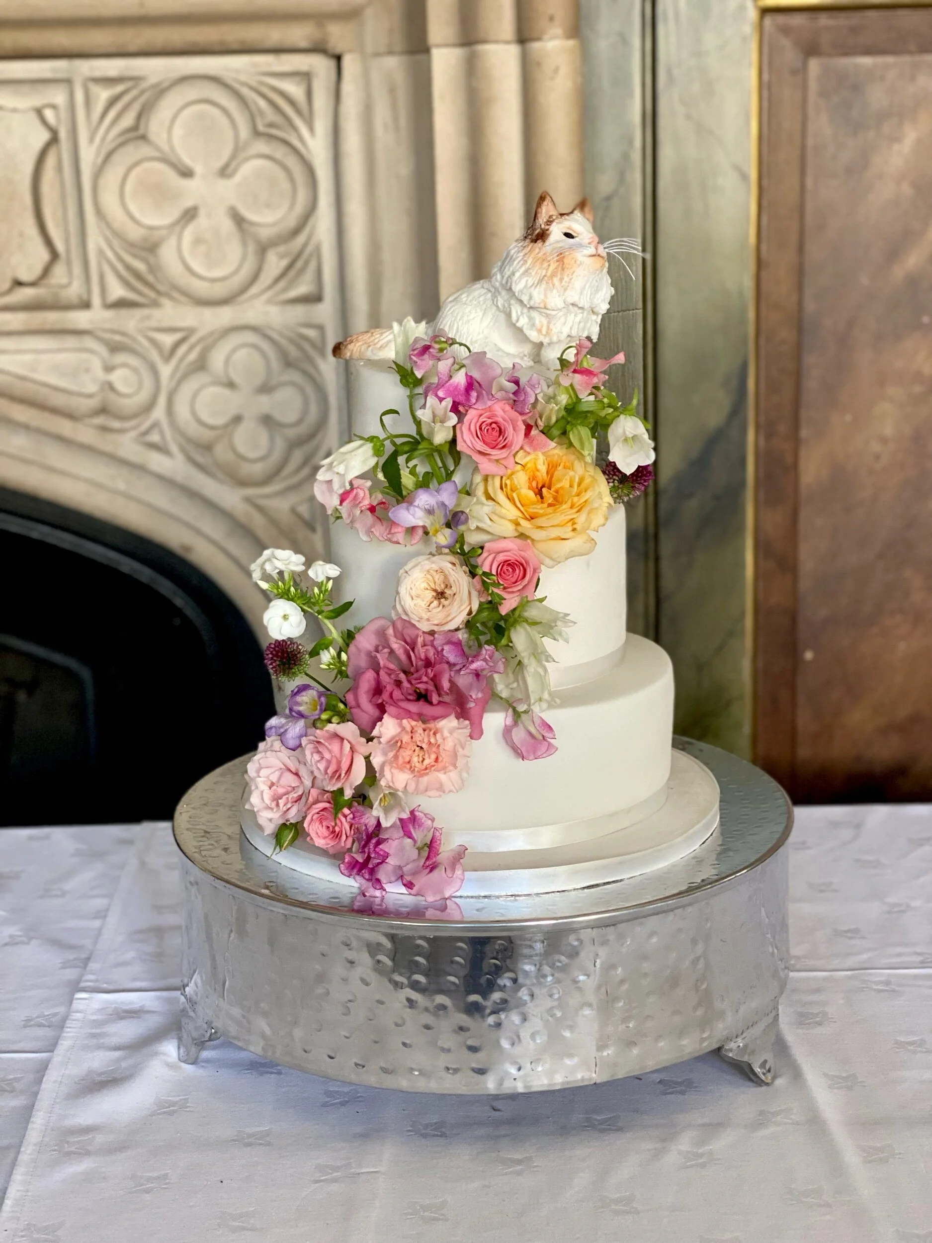 3 tier fondant wedding cake with fresh flowers cascading down the front of the cake topped with a modelling chocolate cat