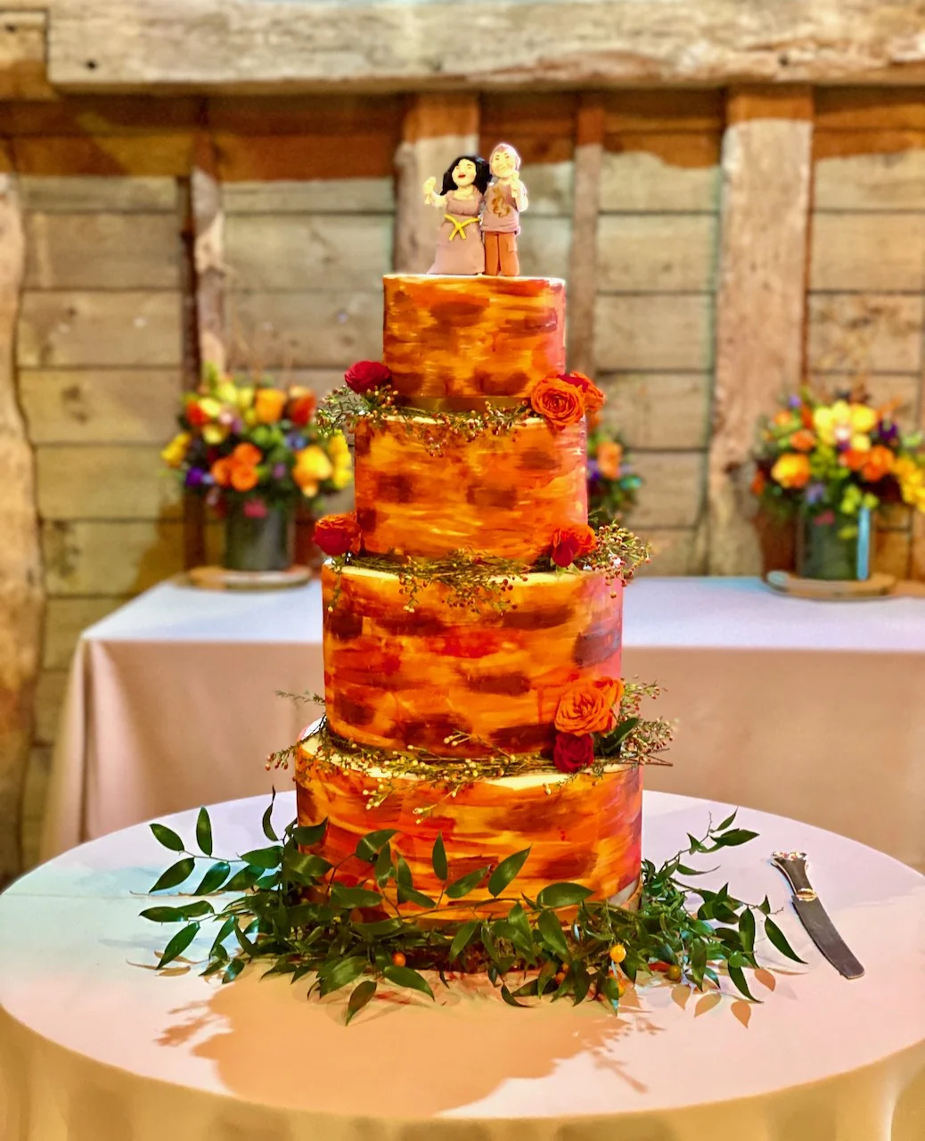 4 tier wedding cake in autumn colours topped with models of the bride and groom. Accents of gold, burgundy, burnt orange and red