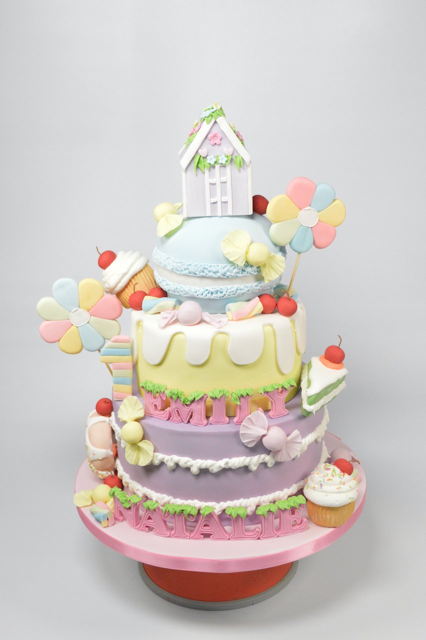 Colorful tiered birthday cake decorated with candies, flowers, and a cottage topper, with the name 'Emily Natalie' written on the bottom tier.