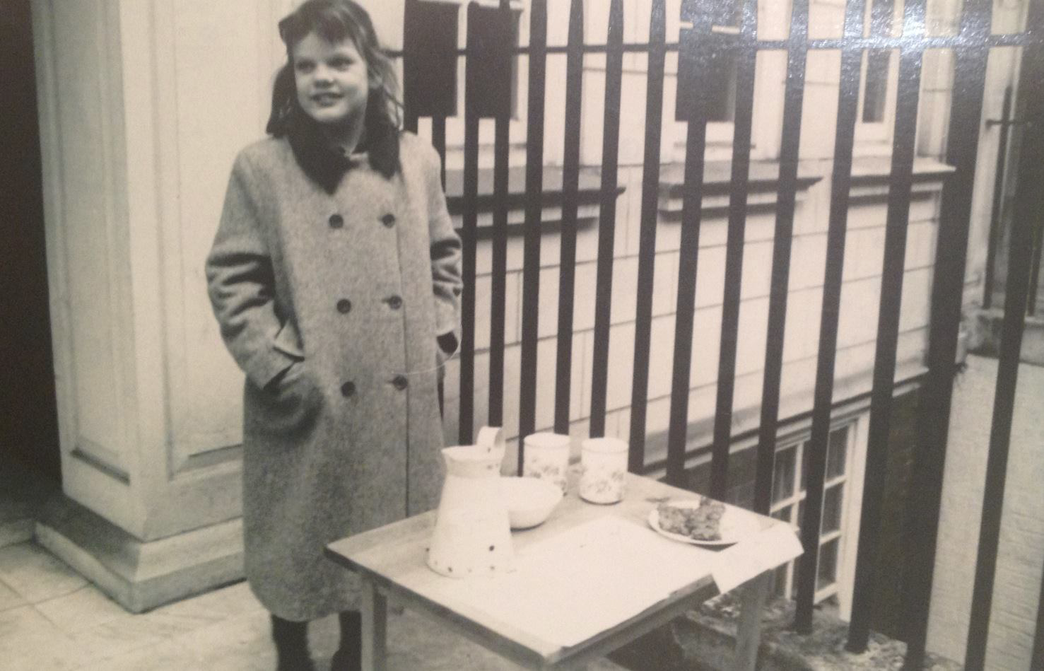 A young girl with dark hair and a coat standing beside a small table with food and drink on it, outdoors in front of a fence and building.