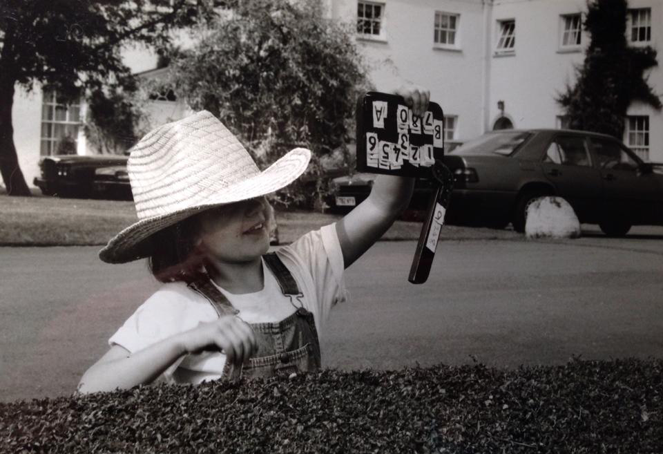 Child wearing a large straw hat and overalls, holding a toy and smiling outdoors in a residential area.
