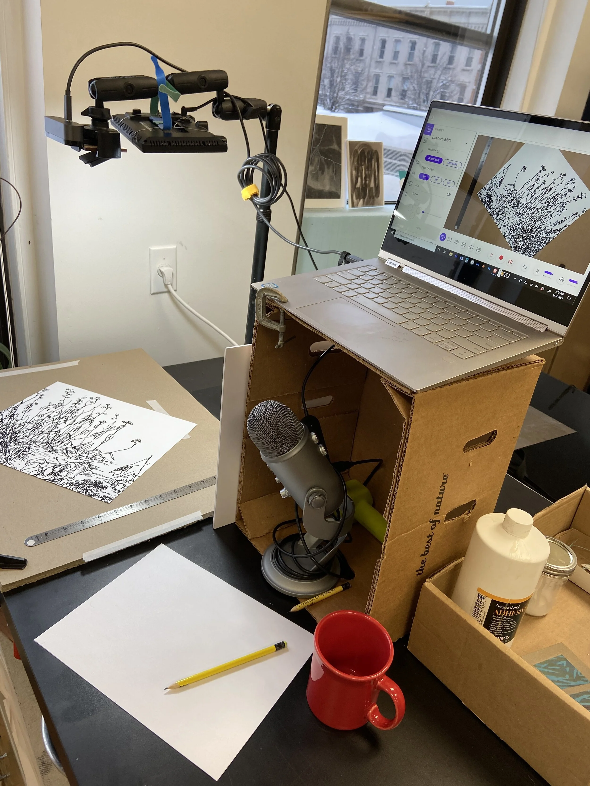 DIY set up for online teaching. Laptop raised up on a tomato box with podcast mic inside. Webcam set up for drawing demo with art materials and cup of coffee.