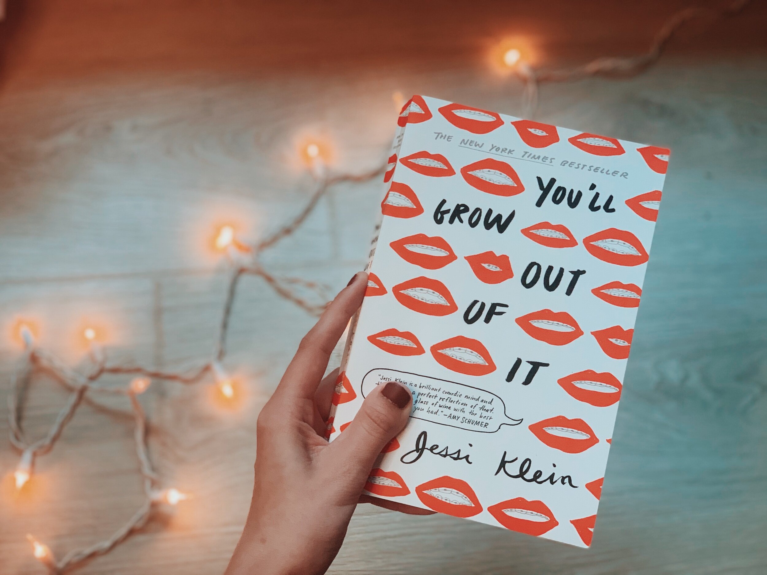 The book You’ll Grow Out of It by Jessi Klein is held up against a light wooden background with a strand of warm twinkly lights in the background it.