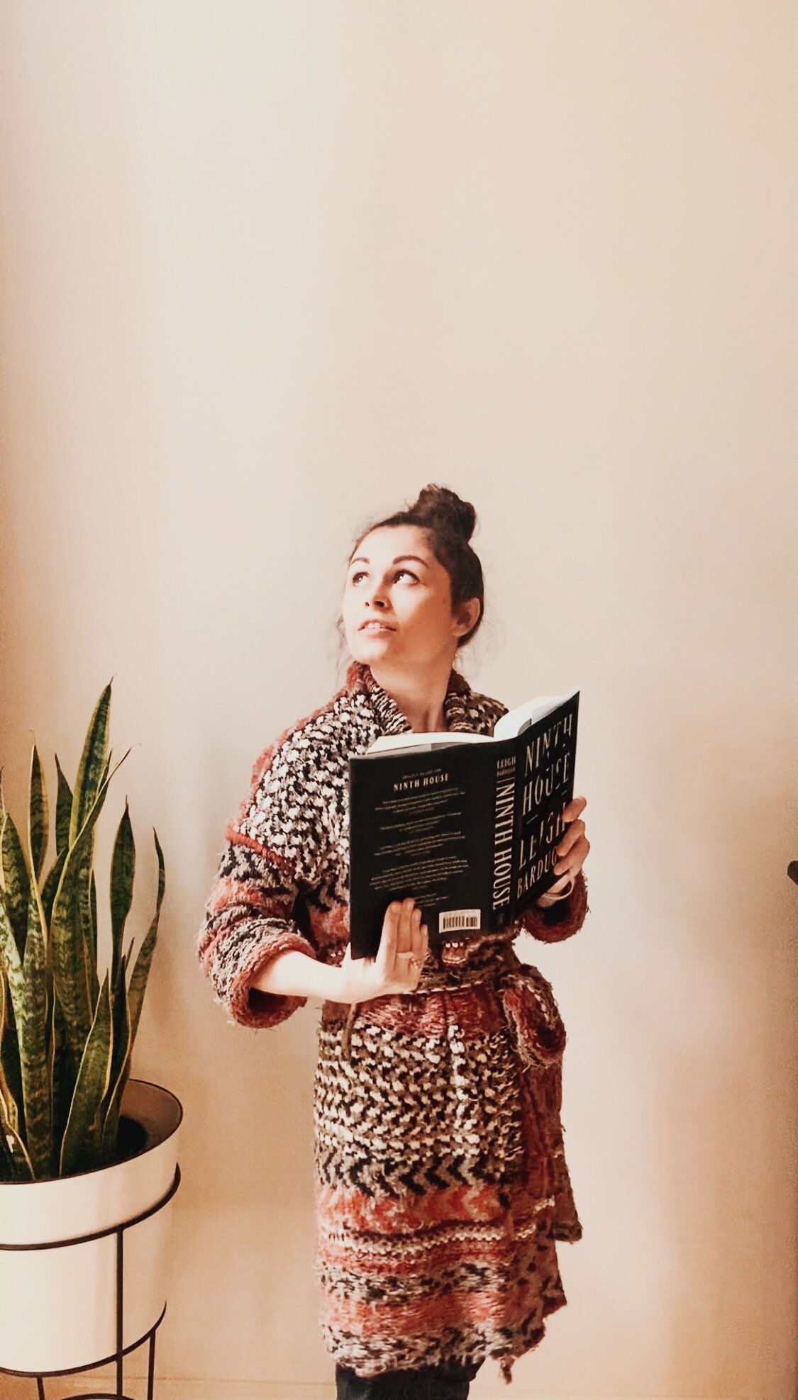 Grace stands in a red sweater holding the book Ninth House by Leigh Bardugo while looking off through the window.