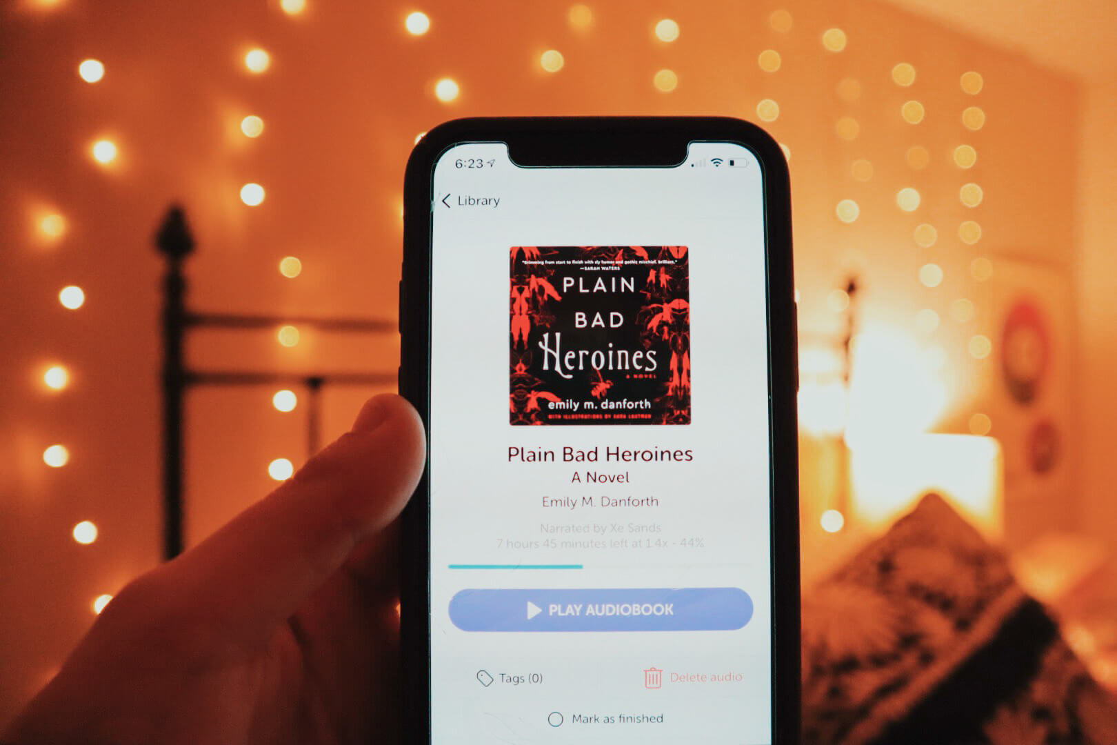 The book Plain Bad Heroines by Emily M. Danforth is featured on an iPhone within the Libro.fm audiobook app. The phone is held up against a warm bedroom background with bokeh lights.