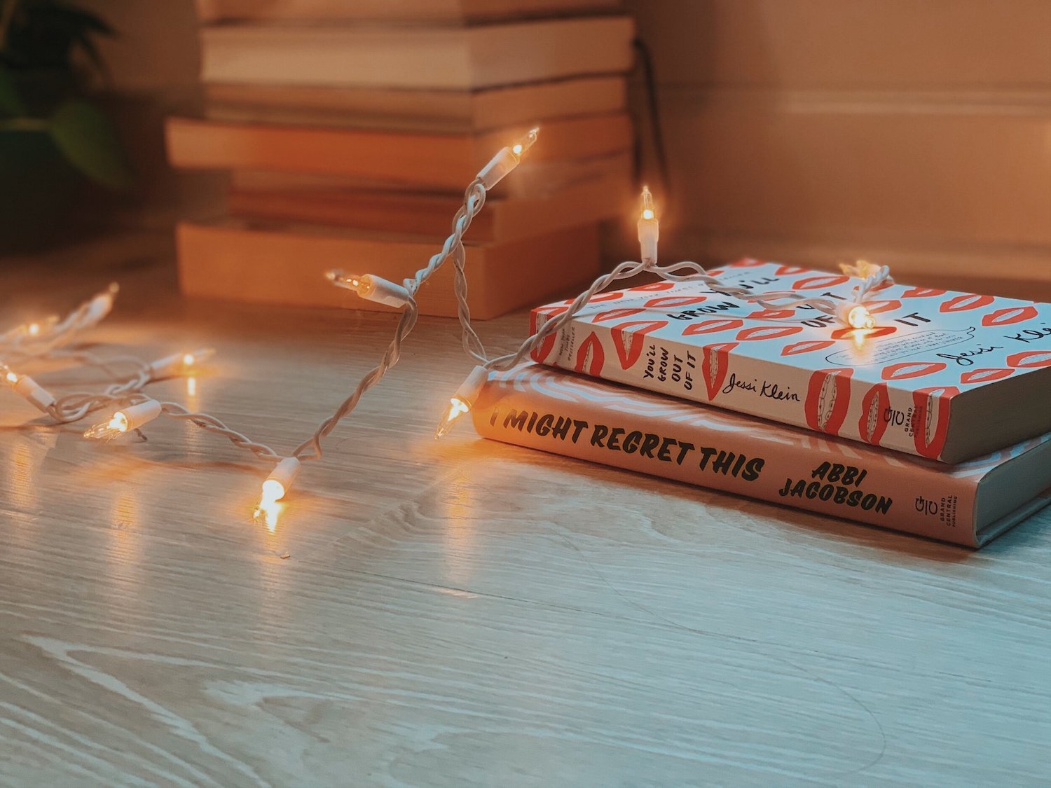 The books You’ll Grow Out of It by Jessi Klein and I Might Regret This by Abbi Jacobson are stacked on a wooden floor next to some other books. A strand of warm twinkly lights lies across them, and there is a potted plant in the background.