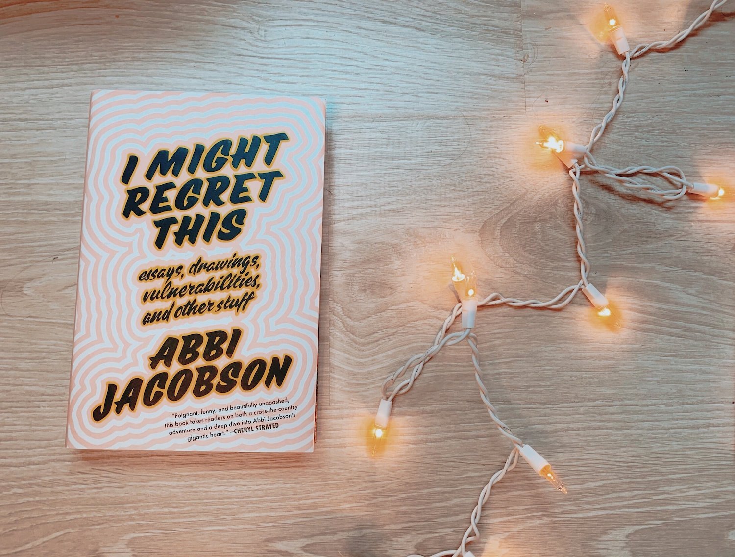 The book I Might Regret This by Abbi Jacobson is against a light wooden background with a strand of warm twinkly beside it.