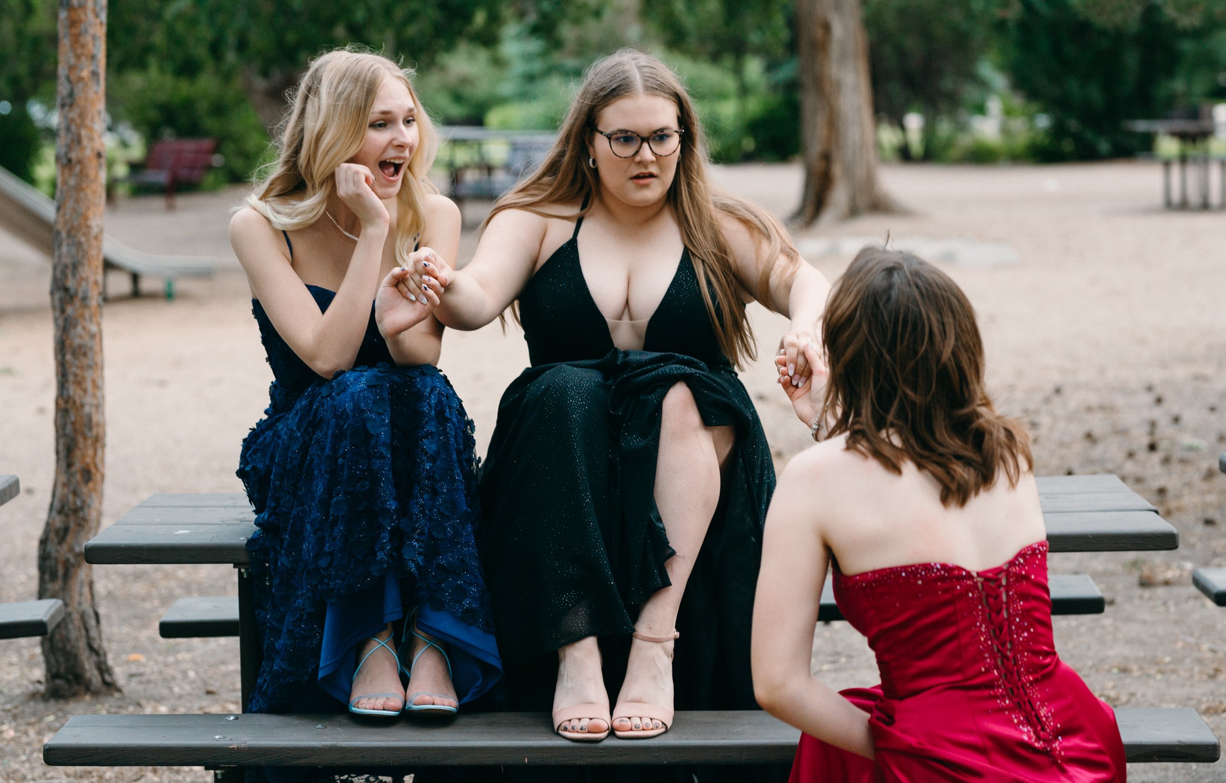  two girls in grad dresses sit on picnic table while the third is down on one knee pretending to propose to the middle girl. her shocked face a funny sight 
