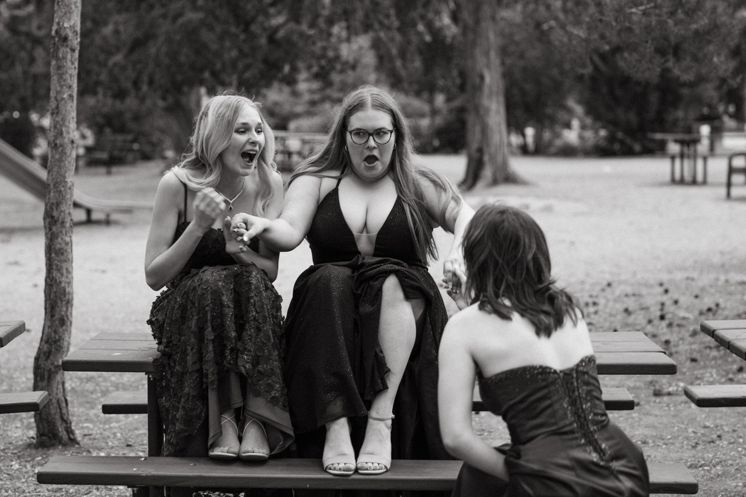  black and white photo of two girls in grad dresses sit on picnic table while the thrid is down on one knee pretending to propose to the middle girl her shocked face a funny sight 