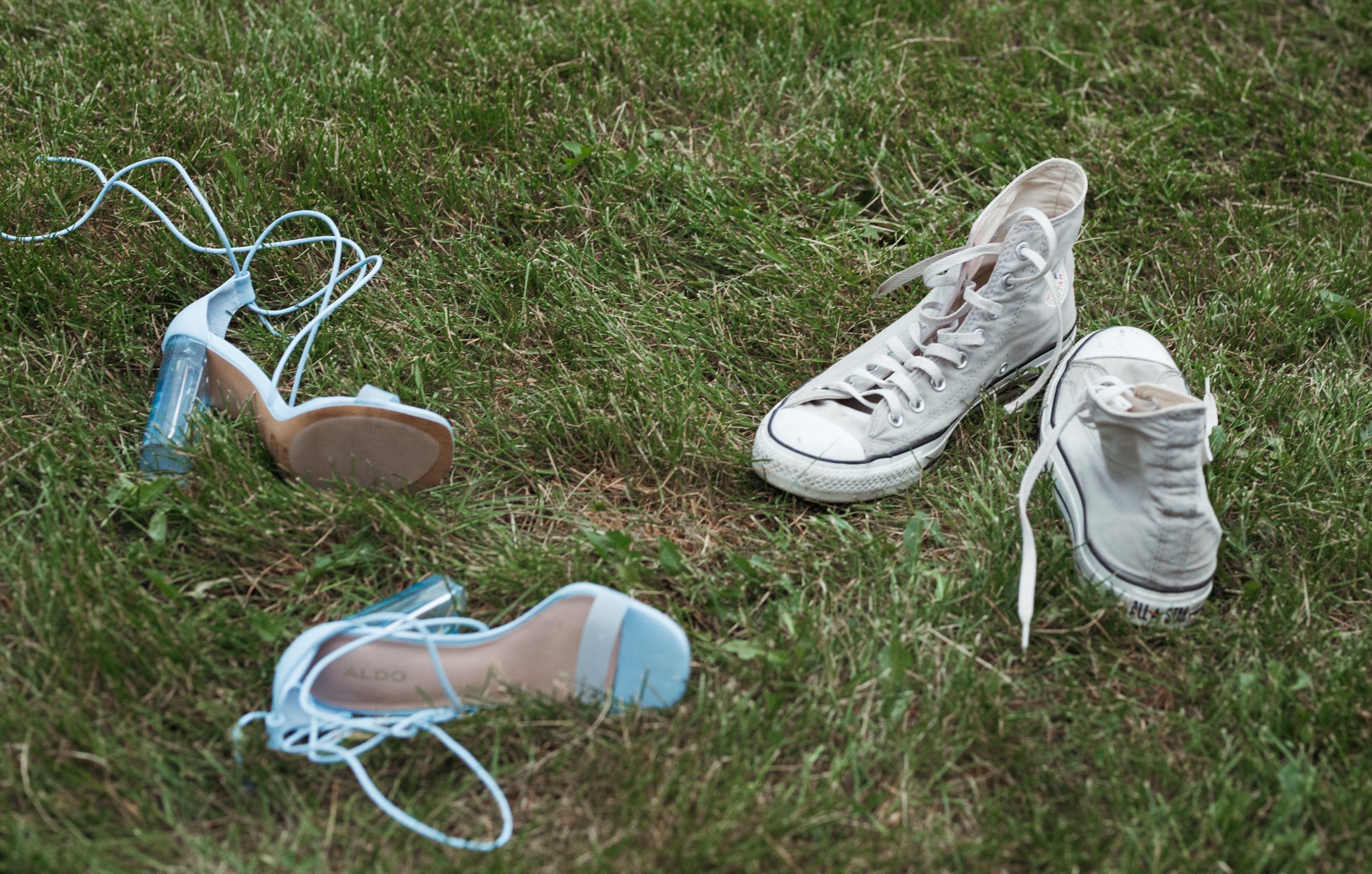  Baby blue strappy high heel shoes and a pair of mens high top sneakers lay  strewn in the grass. 