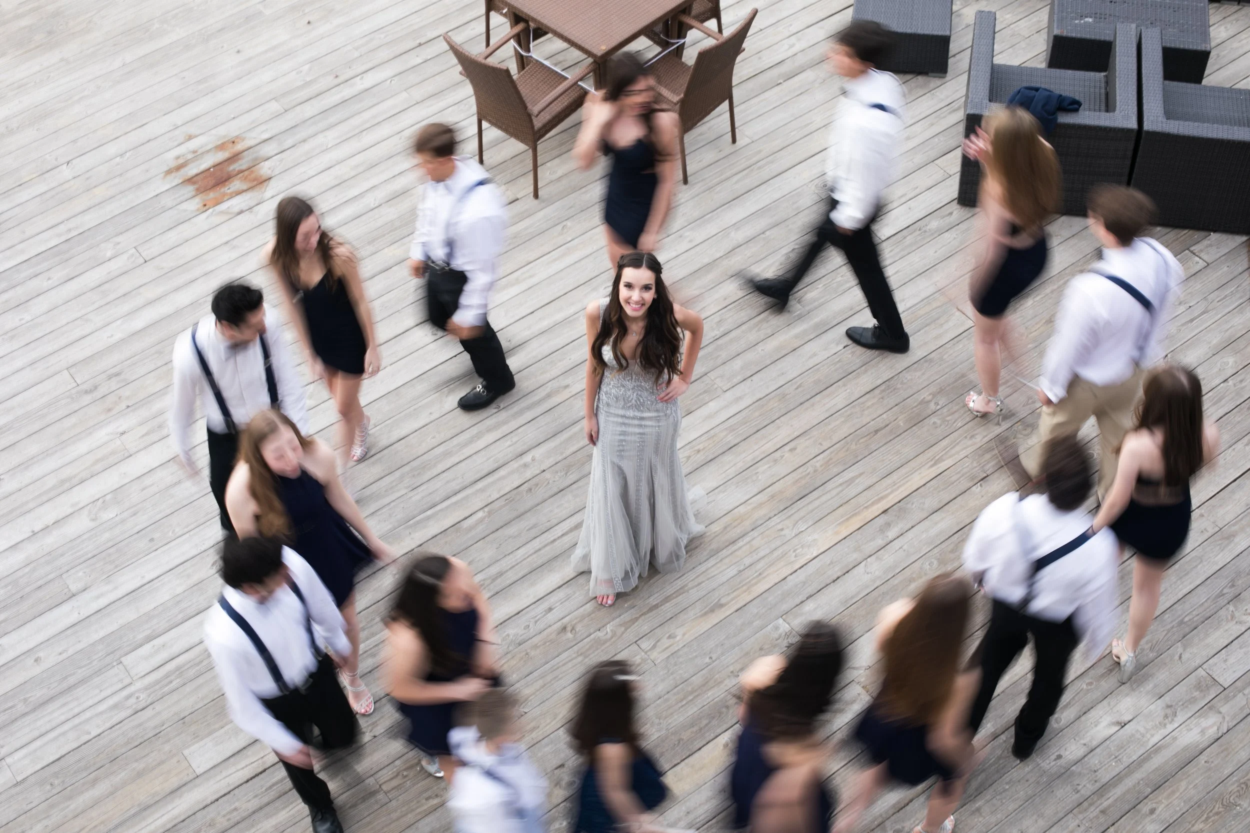 A woman in a silver gown standing on a wooden deck surrounded by people in formal attire. The woman is smiling and looking up at the camera.