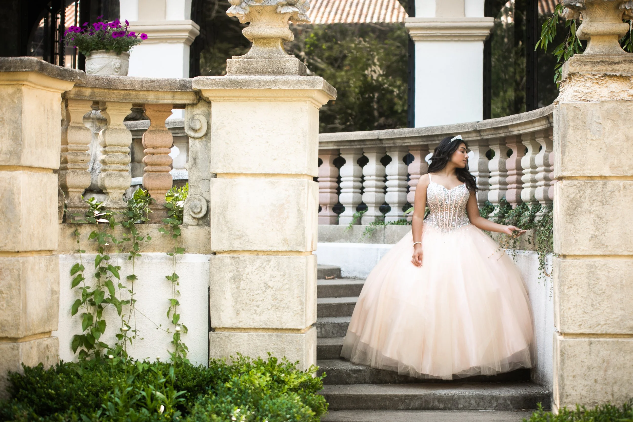 A woman in a light pink ball gown with intricate silver details and a tiara, standing on steps next to a stone balustrade with greenery and purple flowers nearby.