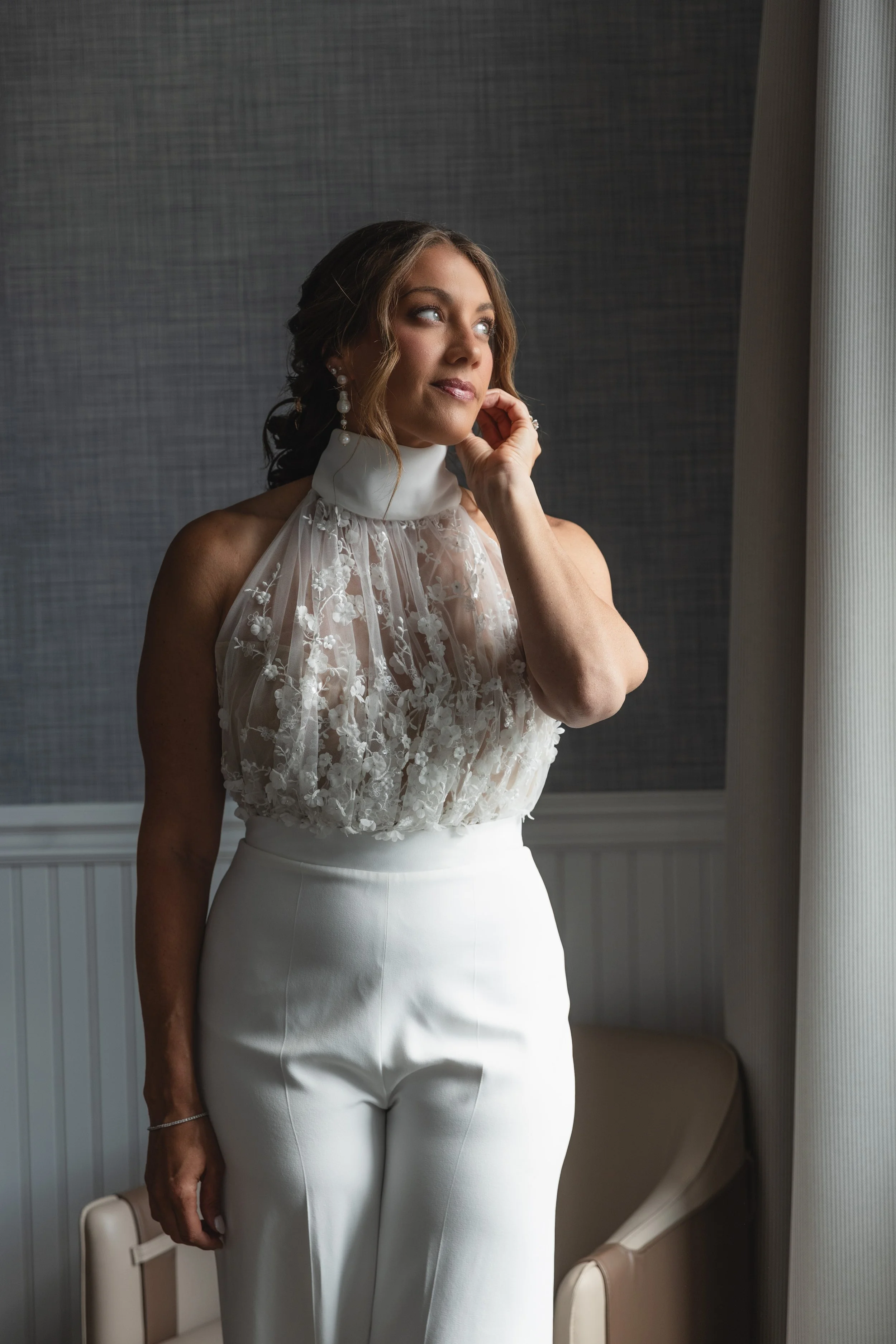 A woman in a white dress with lace details on the top, standing indoors near a window, holding a clutch, with brunette hair styled in loose waves, wearing pearl earrings and a bracelet, gazing thoughtfully, with a textured gray wall behind her.