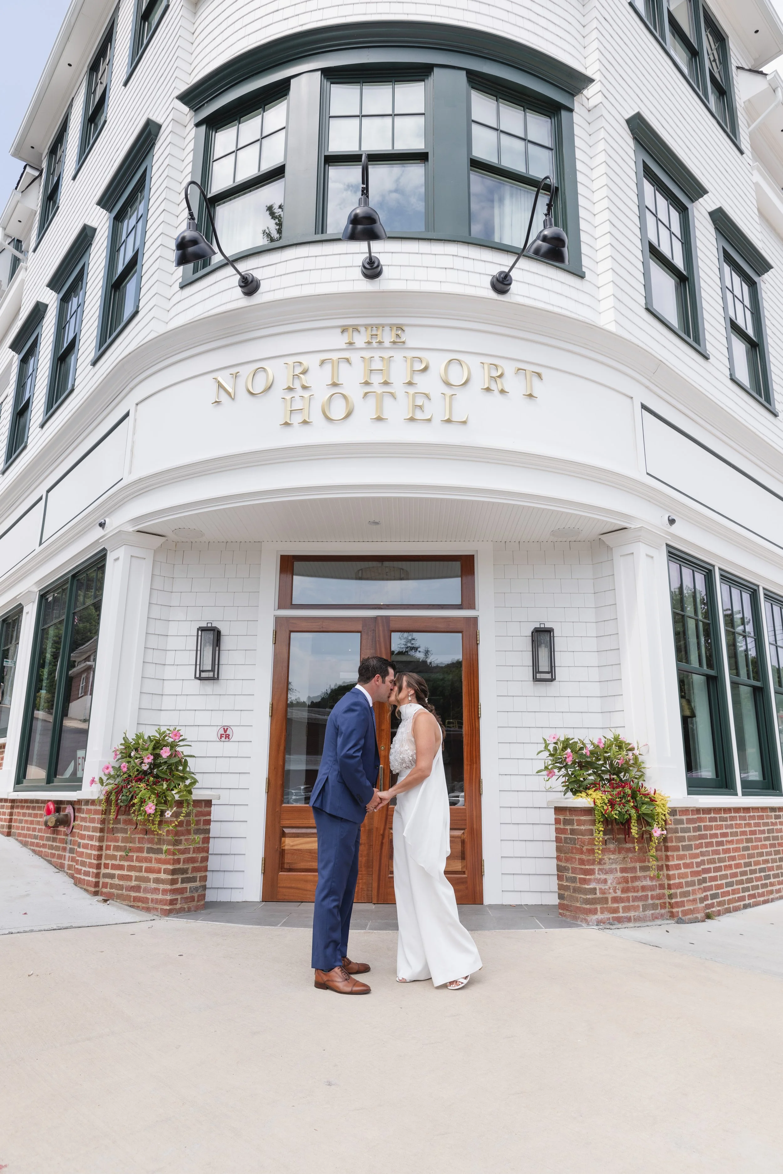 A bride and groom stand close together holding hands in front of a white building with green-framed windows, labeled 'The Northport Hotel,' during their wedding.