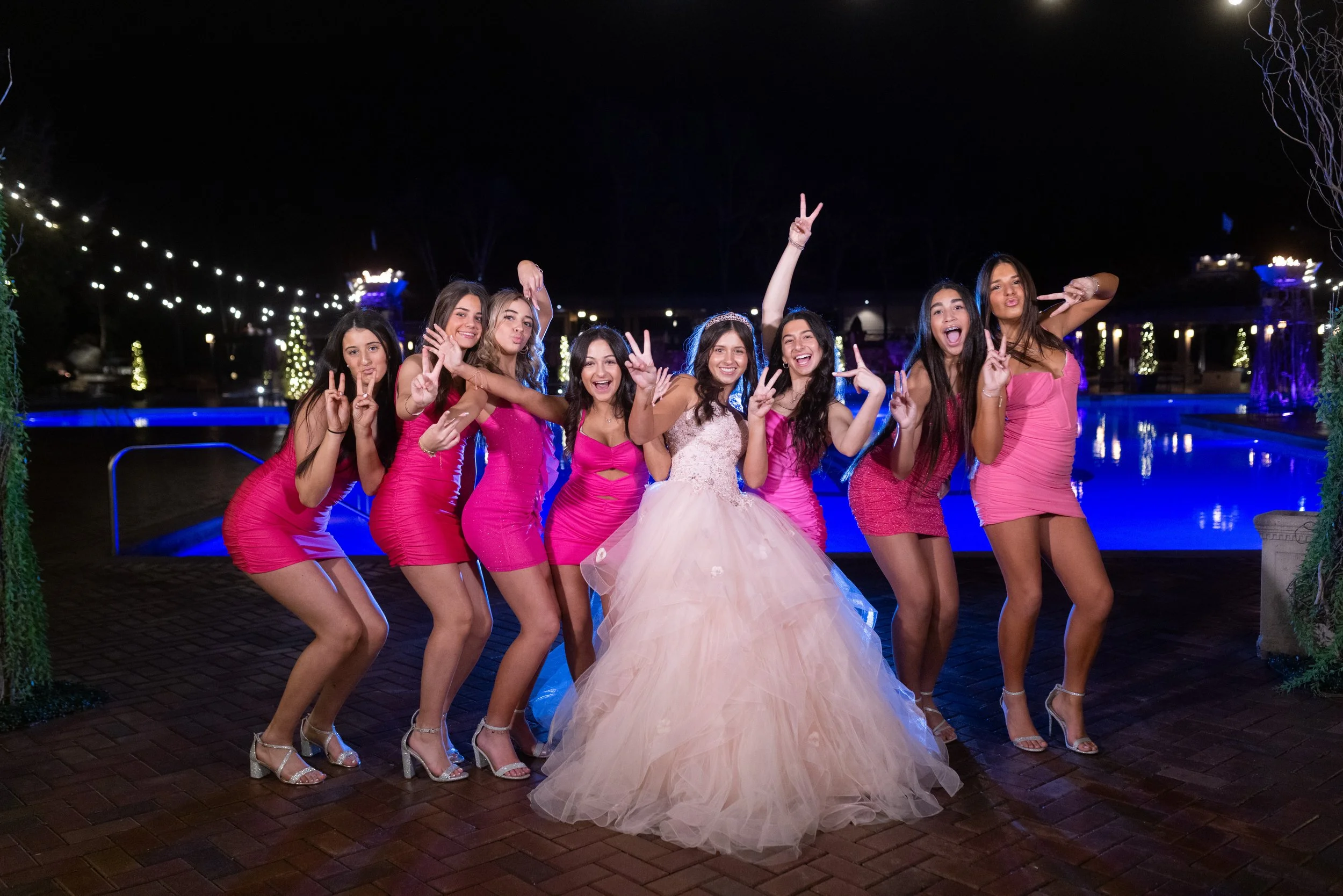 A group of nine young women, dressed in bright pink dresses and high heels, are posing by a pool at night. They are smiling, making peace signs, and one is blowing kisses. The woman in the center is wearing a large, light pink, voluminous gown. Strin