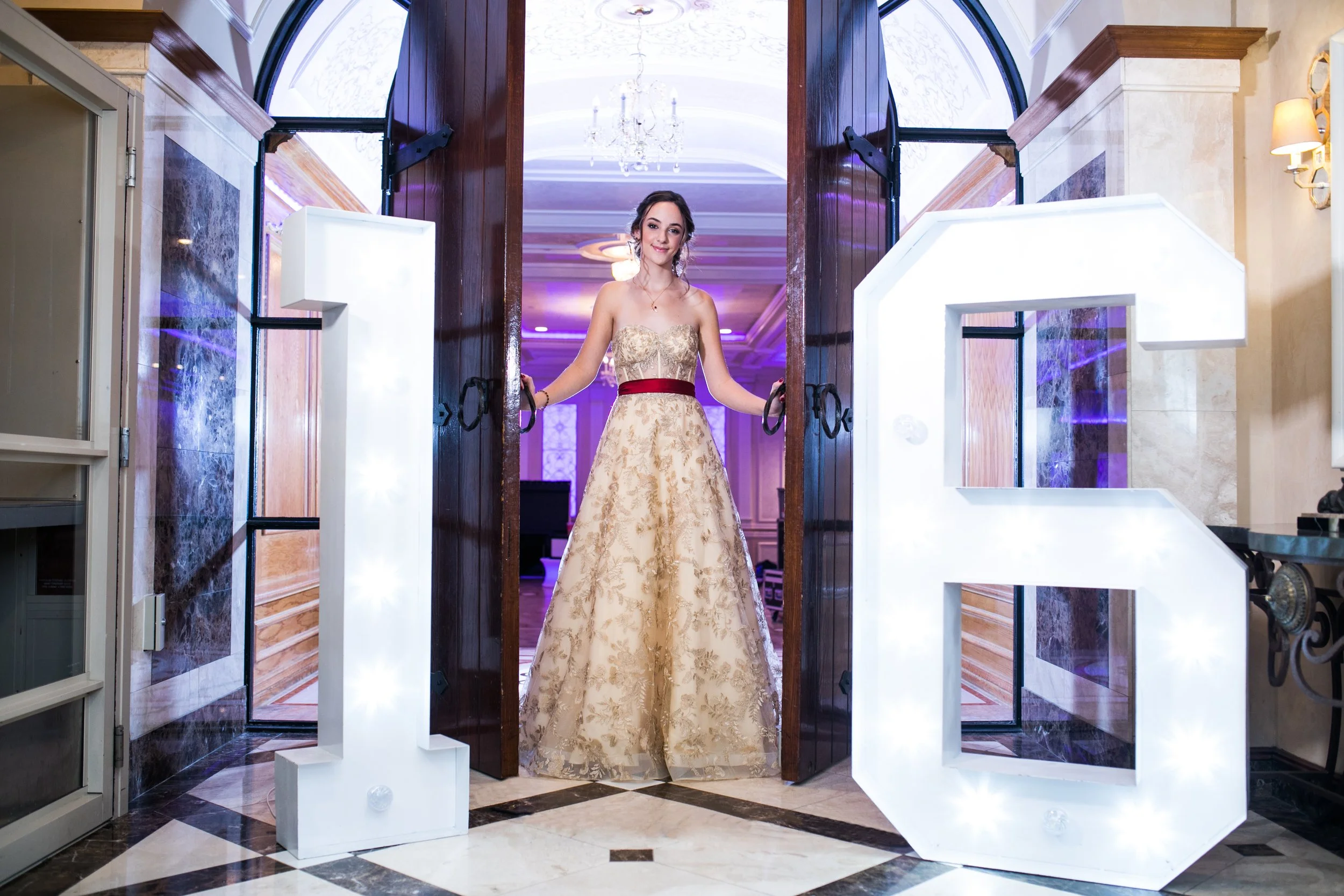 A young woman in a fancy beige and gold dress with a red sash, standing at a doorway, in an elegant decorated room with purple lighting, a chandelier, and wooden accents.