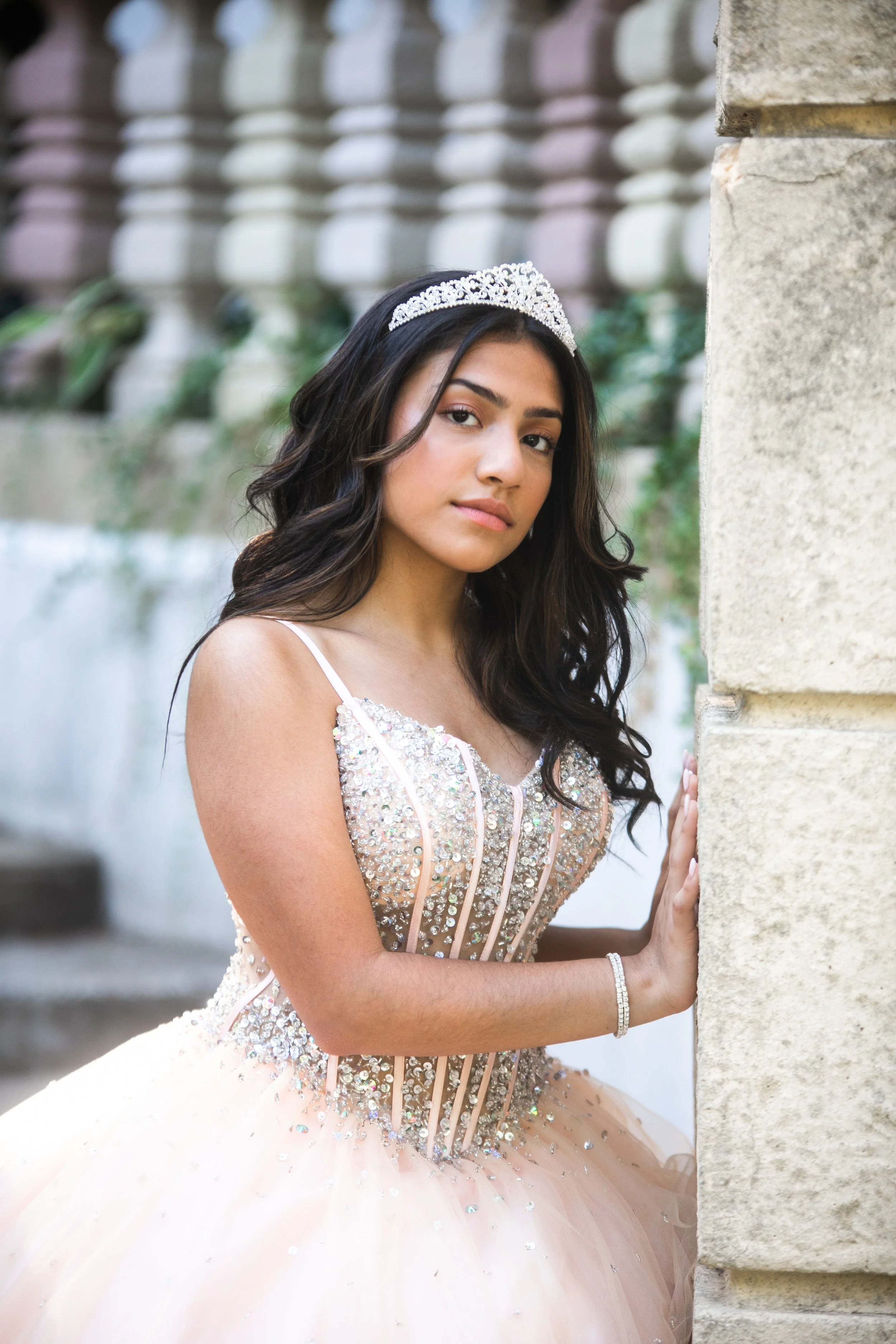 Young woman wearing a tiara and a detailed, sequined pink dress, standing by a stone wall with a blurred background.