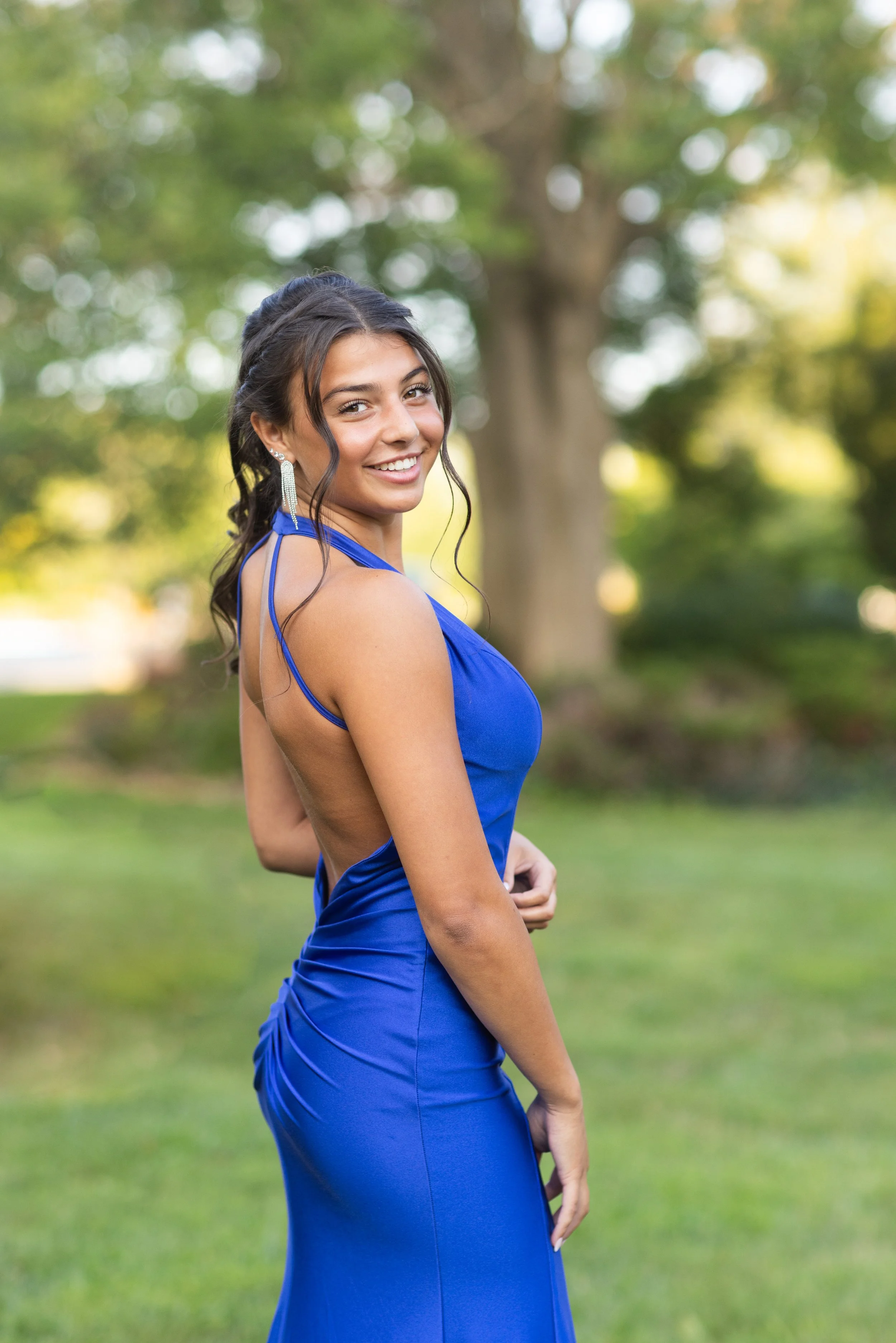 A woman in an elegant royal blue dress with open back and thin straps, smiling in a park with a blurred green background of trees and grass.