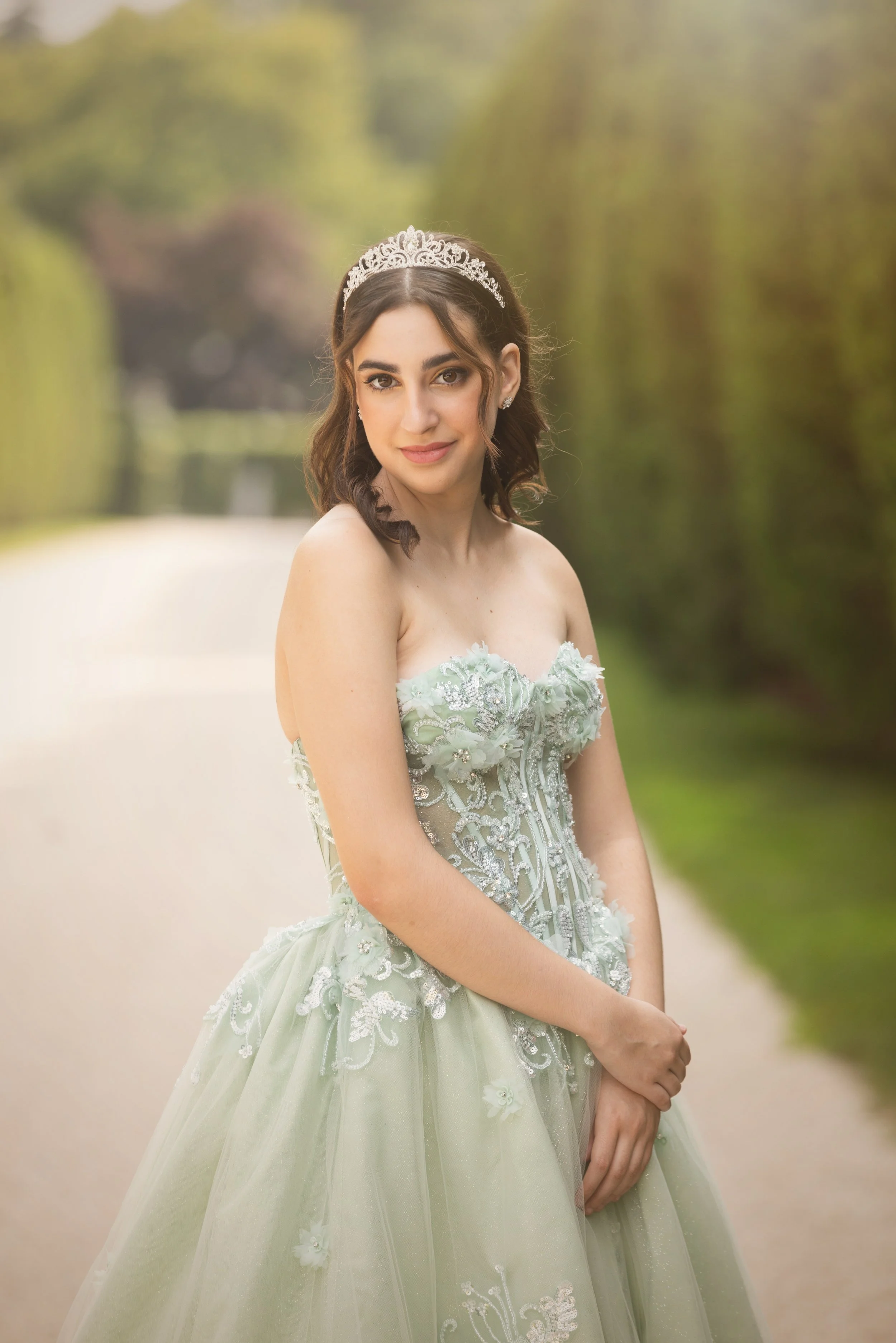 A young woman in a strapless, light green ornate gown with floral embroidery, wearing a tiara and earrings, standing outdoors on a pathway with greenery and trees in the background, looking at the camera with a gentle smile.