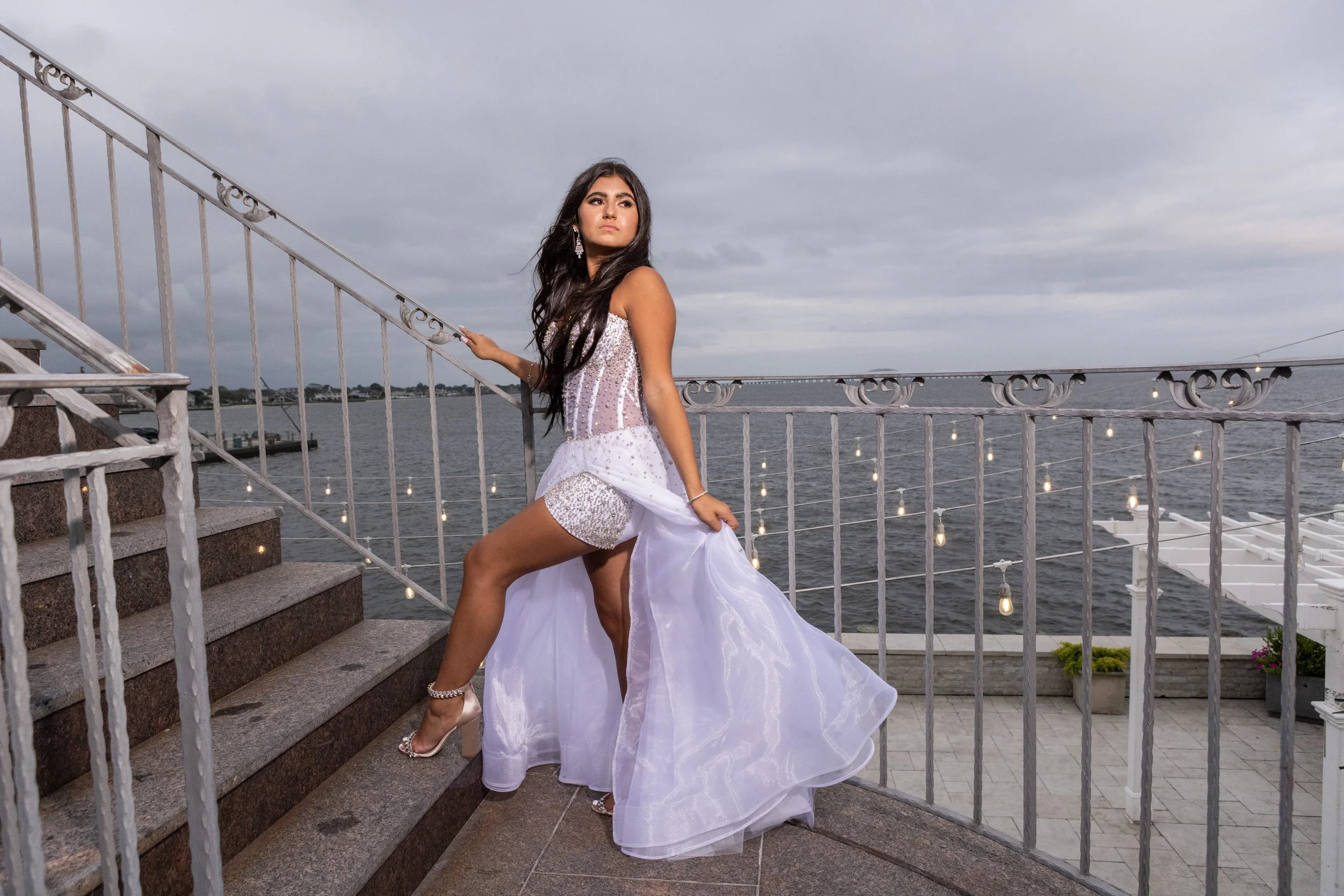 A young woman in a formal gown standing on stairs by the water, posing with a hand on the stair railing and looking to the side, with a cloudy sky and a bridge in the background.