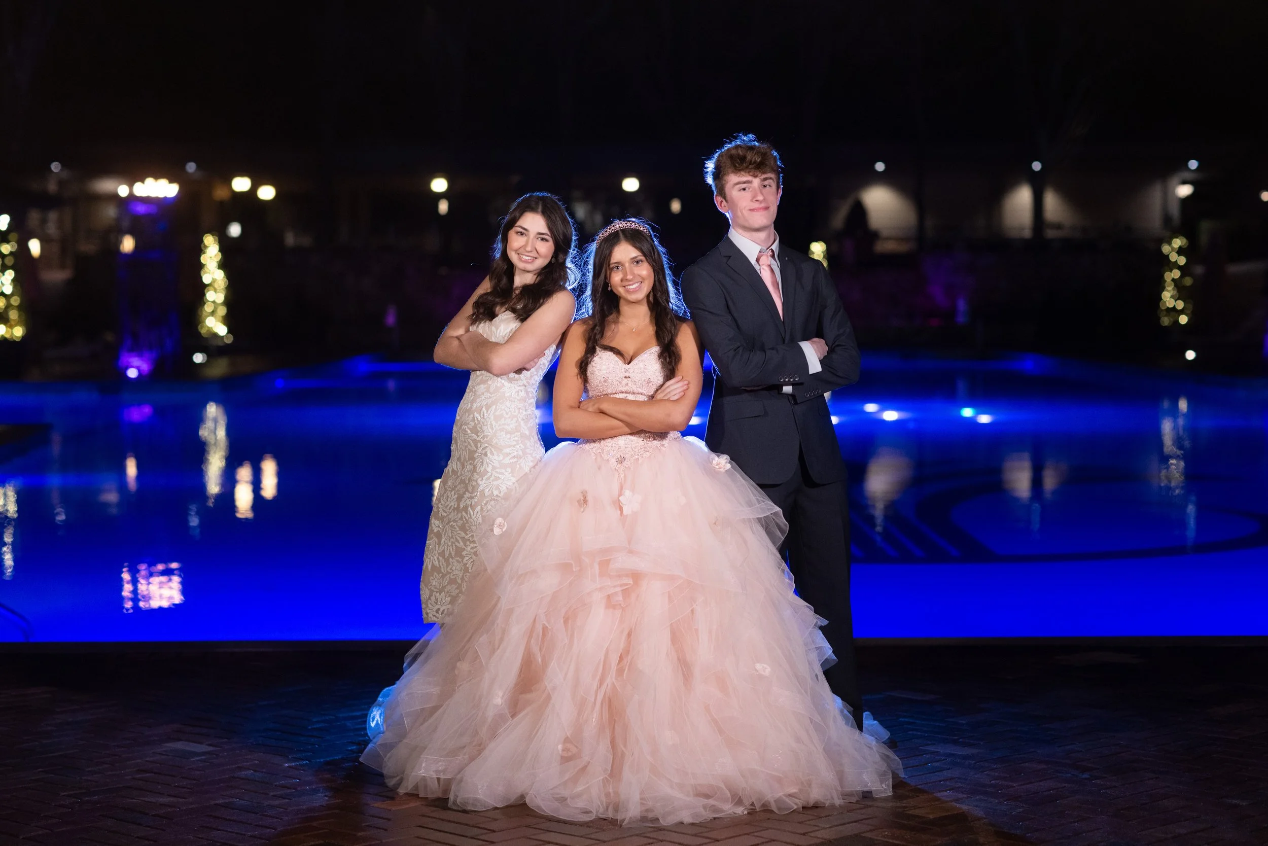 Three teenagers, two girls and one boy, dressed formally at night by a pool with festive lights in the background.