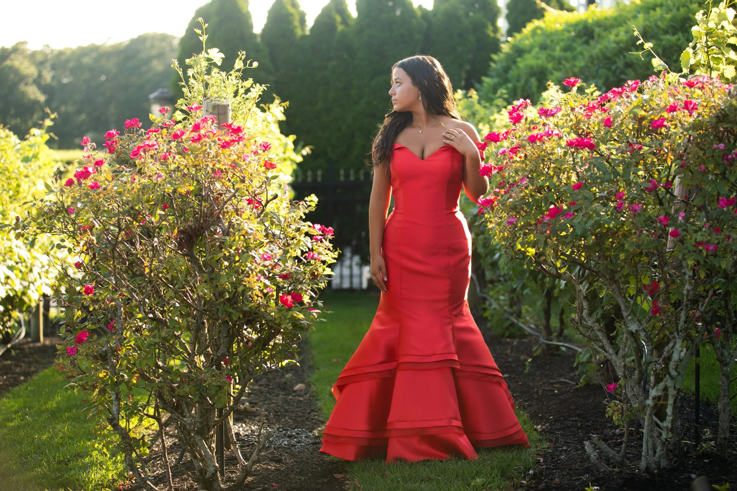 A woman in a red dress standing between flowering bushes in a garden during daytime, looking to her left.