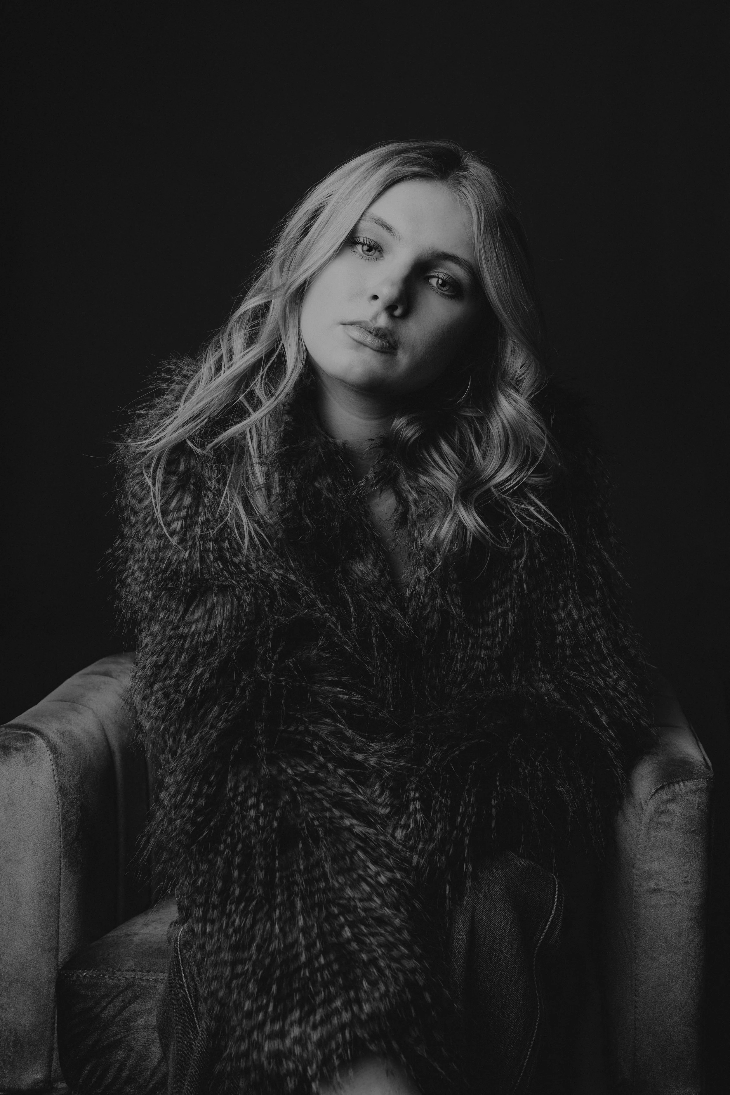 A black and white portrait of a woman with wavy hair, wearing a fur coat, sitting on a couch against a dark background.