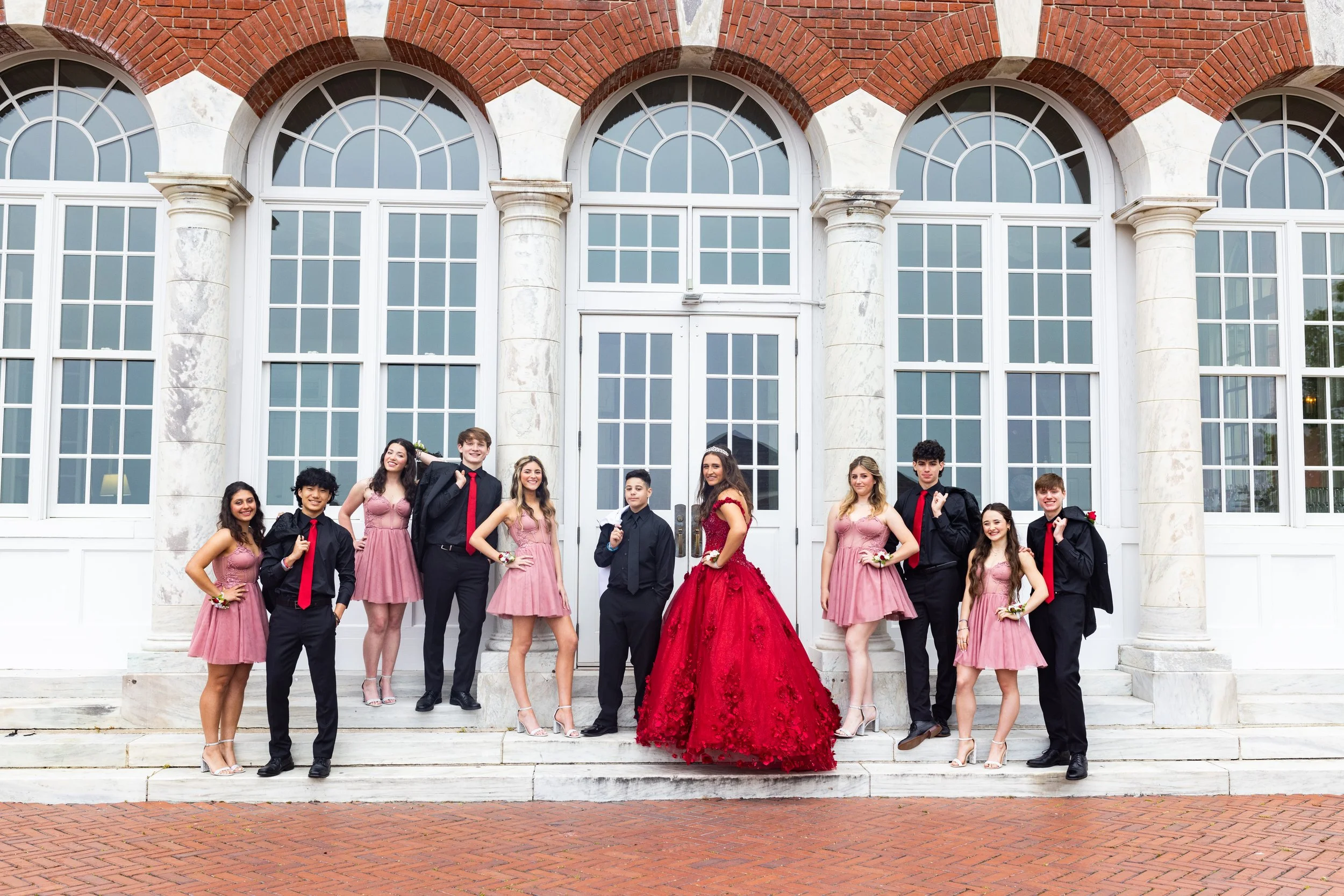 Group of young people dressed in formal wear standing on steps in front of a large building with arched windows and brick facade, celebrating a special occasion.