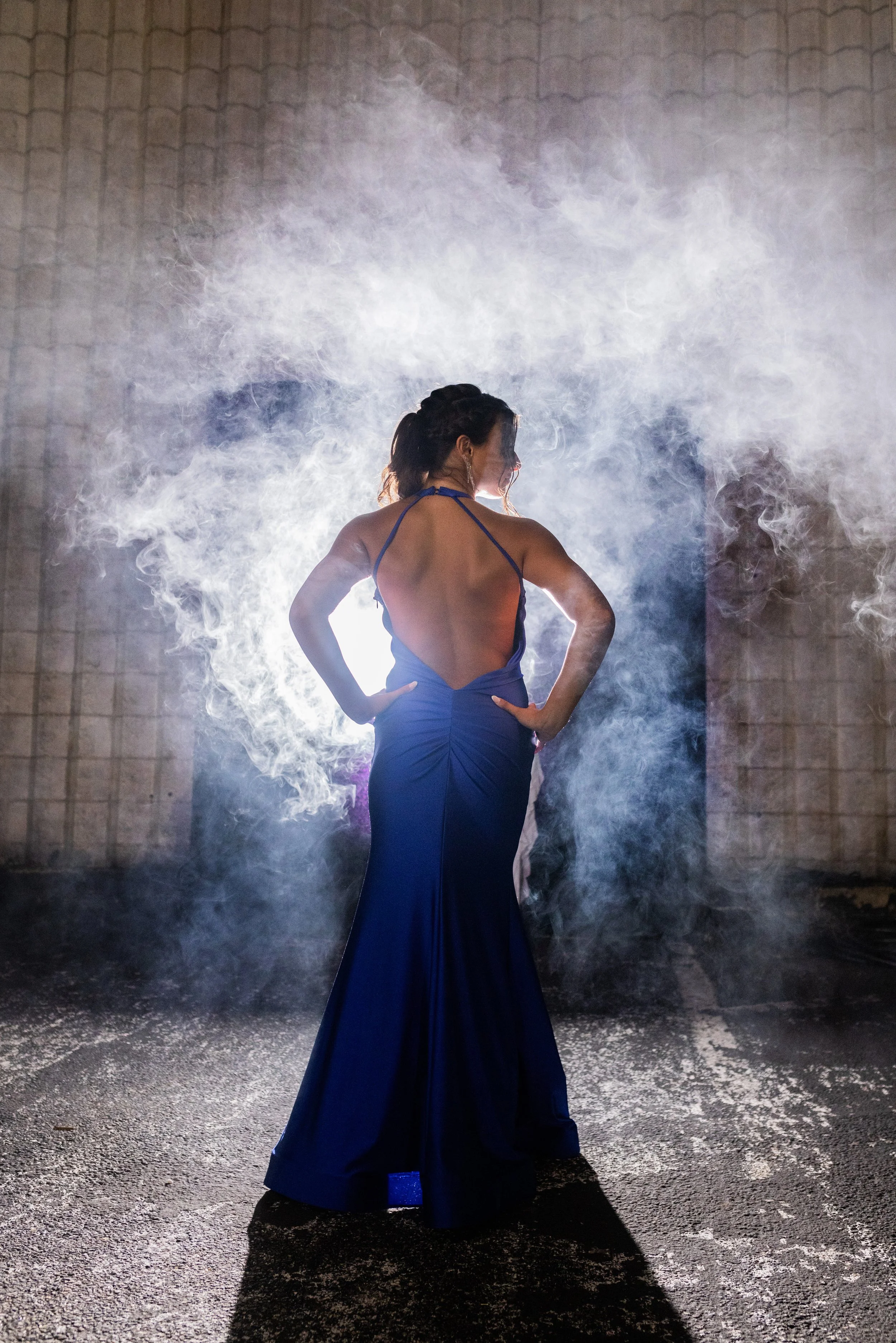 A woman stands in front of a bright light with smoke around her, wearing a dark blue gown with an open back and a halter neckline, posing with her hands on her hips.