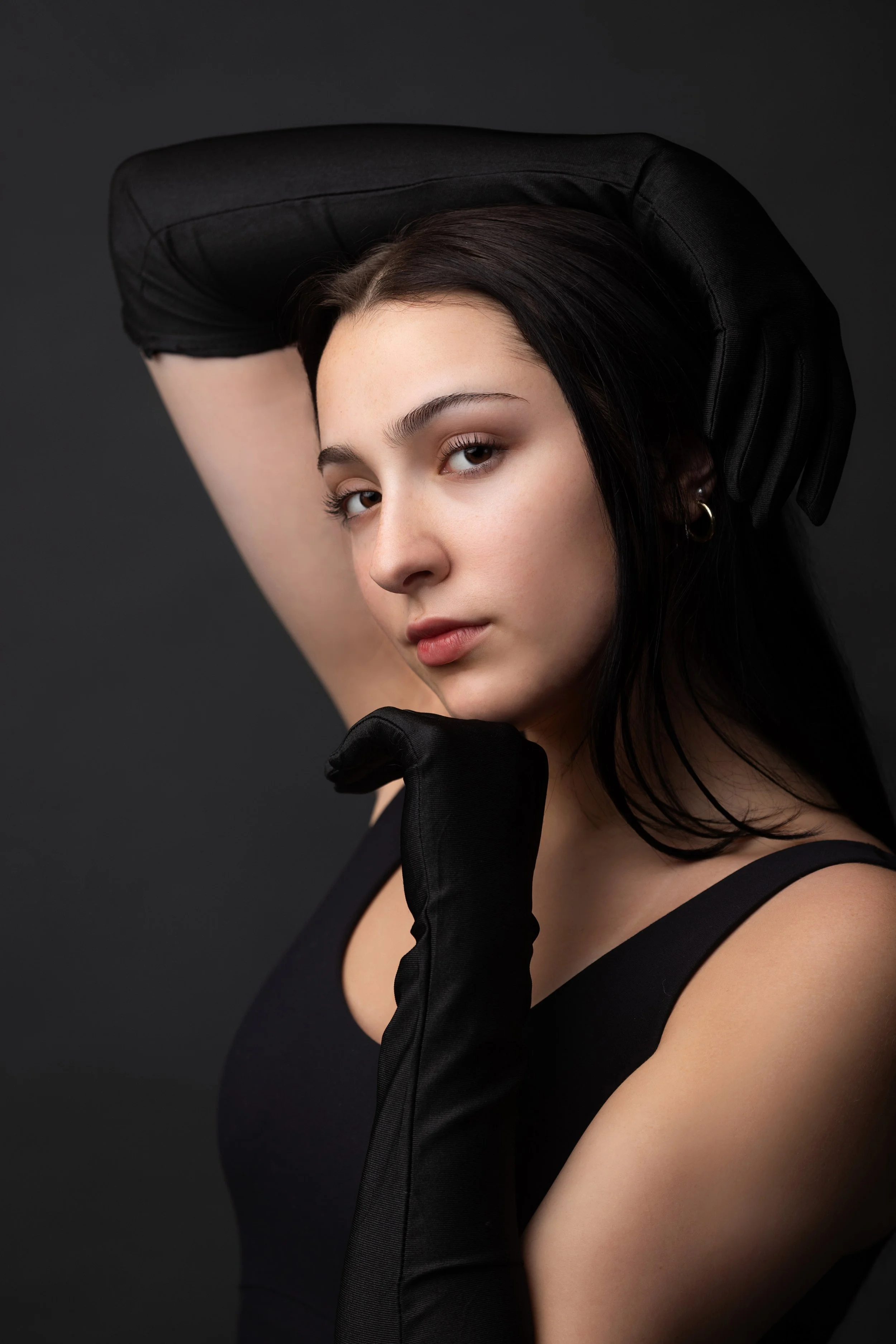 A young woman with dark hair and earrings wearing a black sleeveless top, posing with her left arm raised over her head and her right hand near her face, against a dark background.
