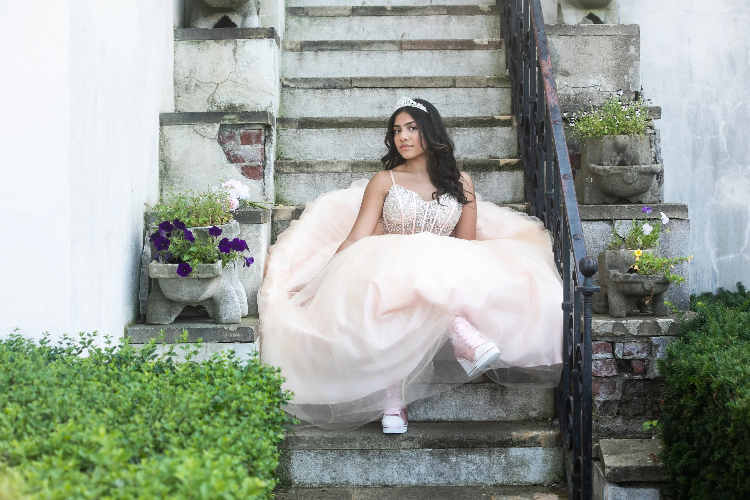 Young woman sitting on outdoor stone steps wearing a glittery pink dress, tiara, and sneakers, surrounded by potted flowers and greenery.