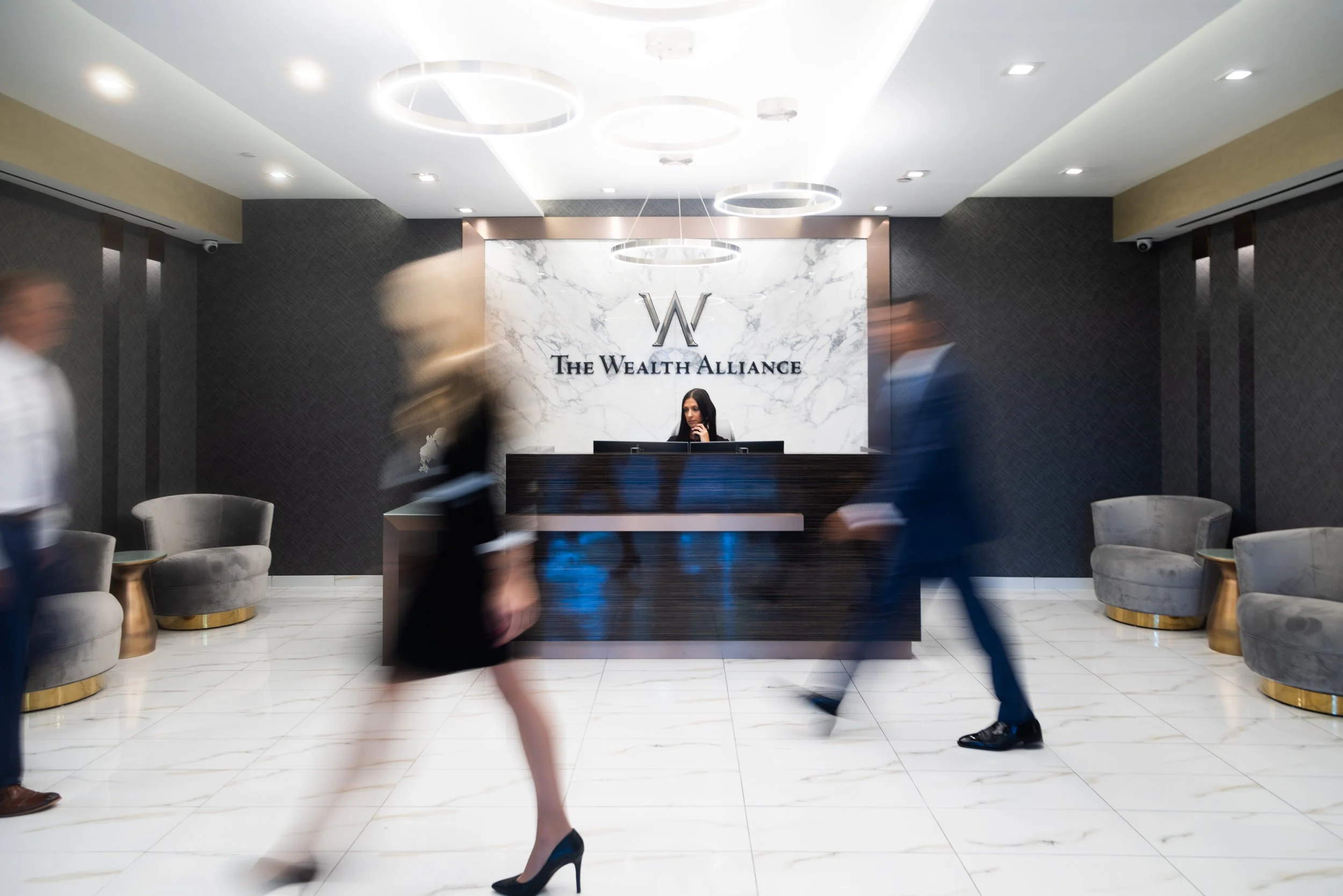 Front desk area of The Wealth Alliance with a marble wall, logo, and seated chairs, people walking past.