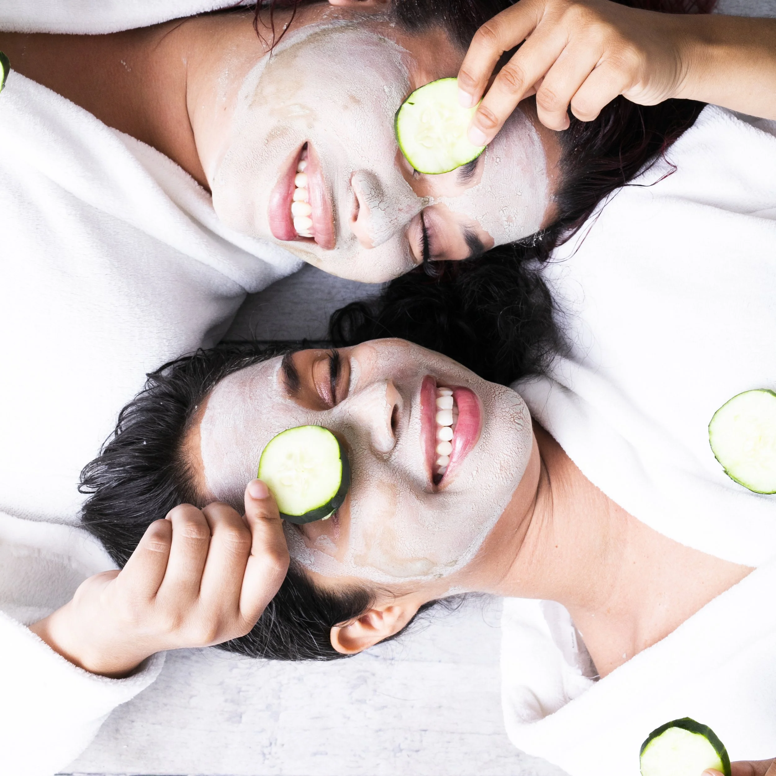 Two women lying down with white face masks, holding cucumber slices over their eyes, smiling.