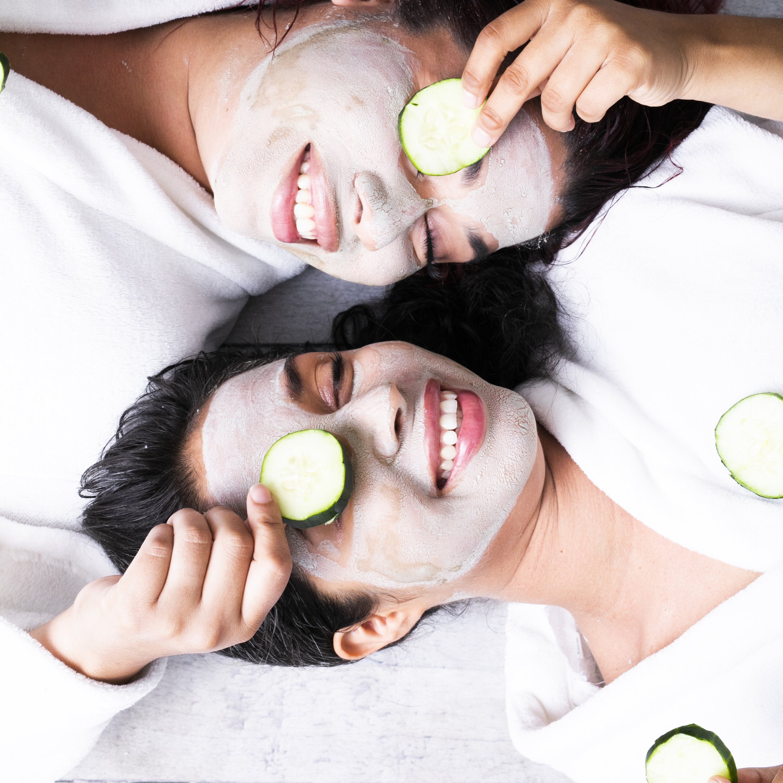 Two women lying down with white face masks, holding cucumber slices over their eyes, smiling.