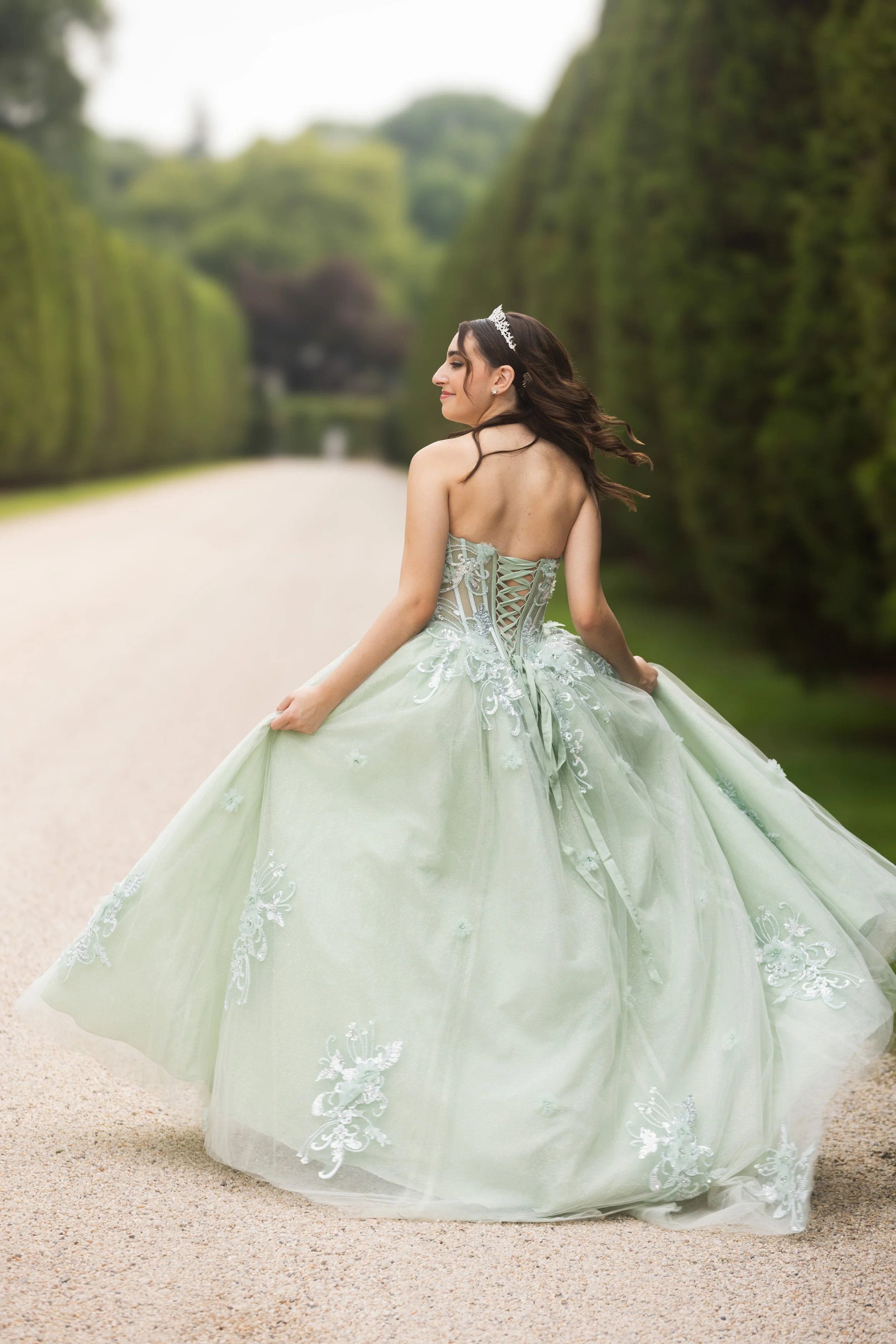 A woman in a pale green wedding gown with lace and embroidery details, wearing a tiara, standing on a gravel path lined with tall, green hedges, with trees and a building in the background.