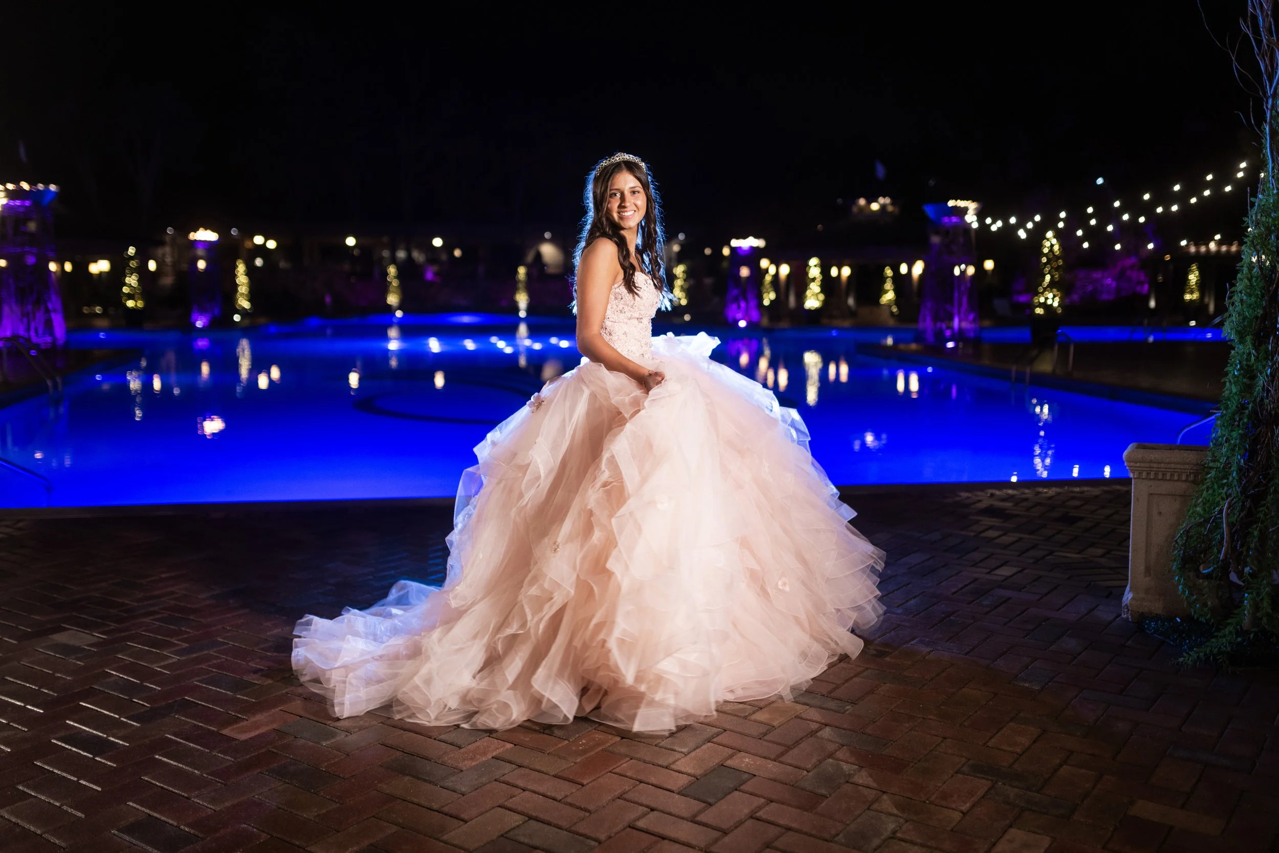 Young woman in a formal gown and tiara standing by a lit swimming pool at night with string lights in the background.