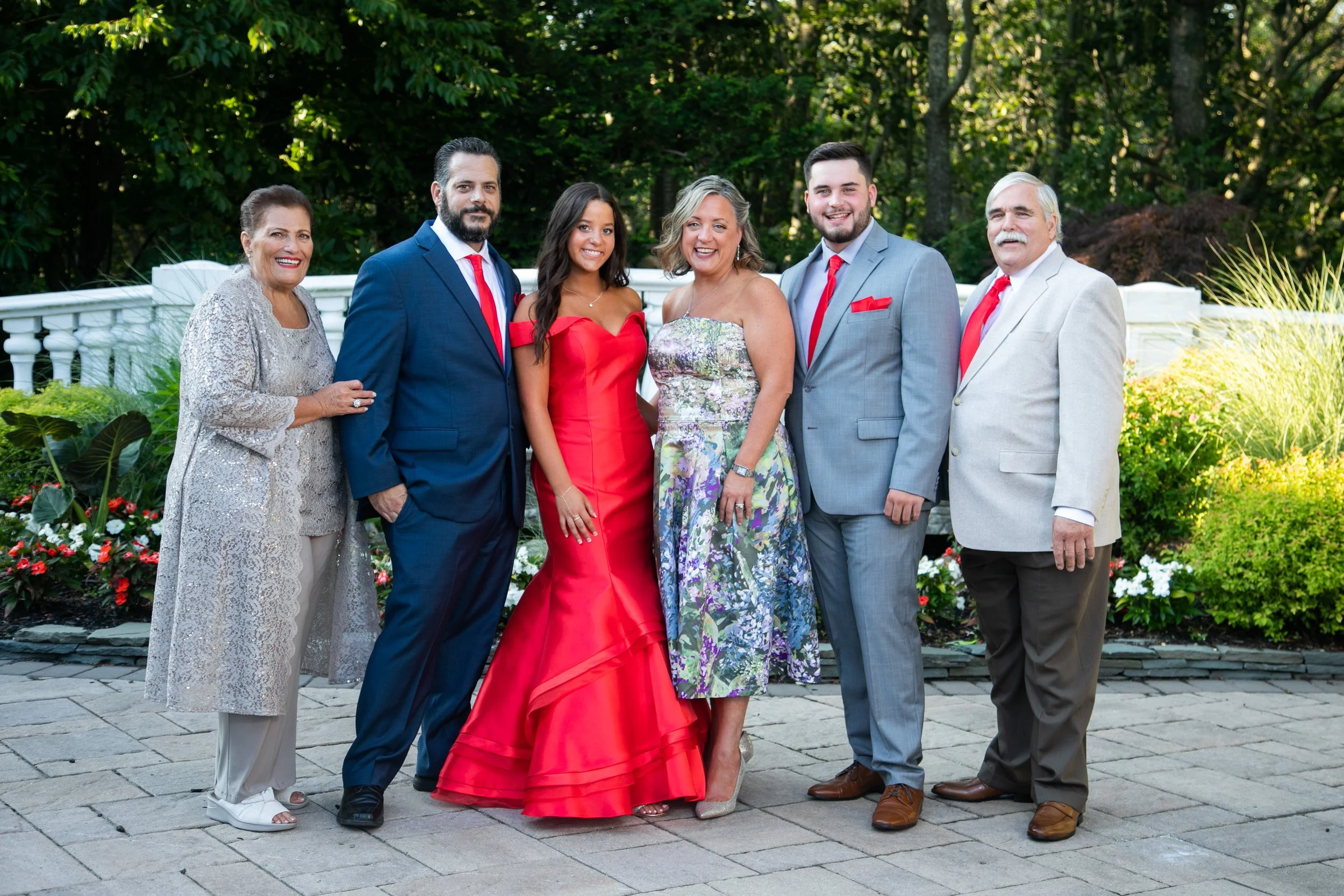 A group of seven people dressed in formal attire, standing outdoors on a paved area with greenery and trees in the background, smiling for a photo.