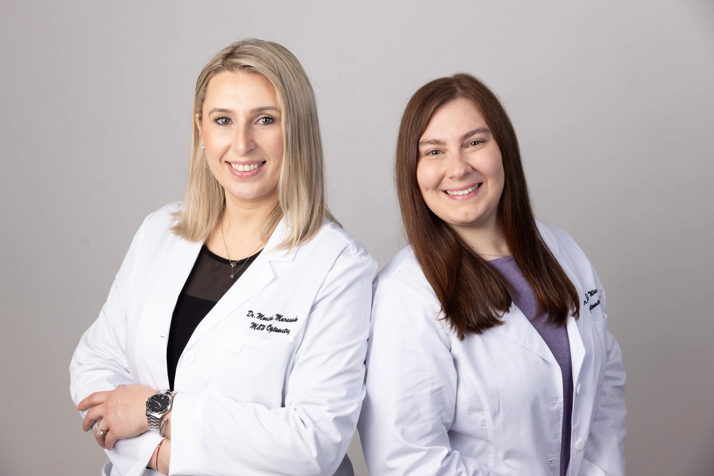 Two women in white lab coats, smiling, standing against a plain gray background. One has blonde hair and wears a watch, the other has brown hair and wears a purple shirt underneath her coat.