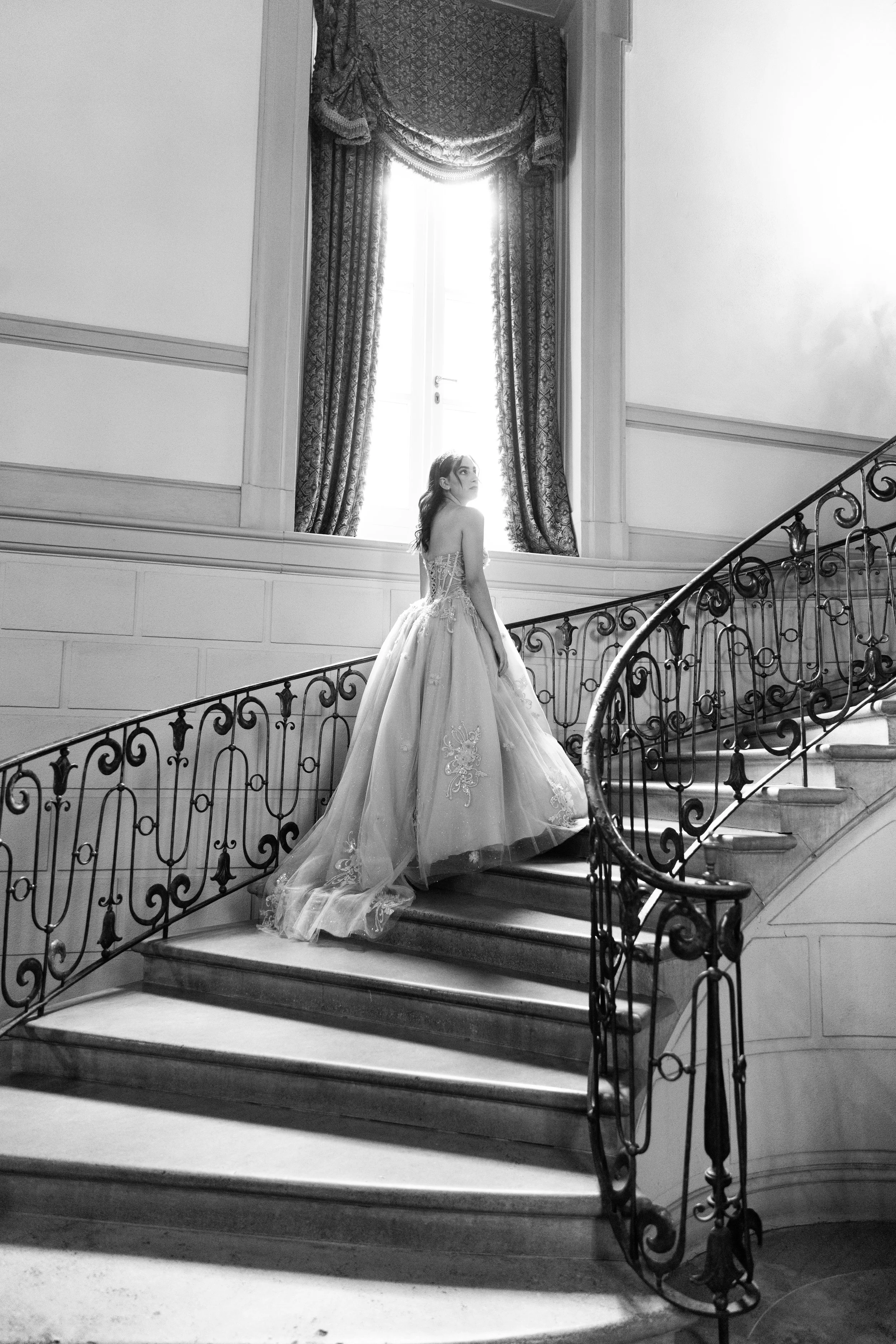 A woman in a wedding dress standing on a grand staircase in an elegant, historic building with tall windows and ornate drapes.